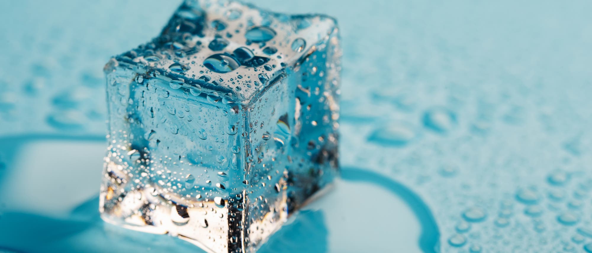 Ein einzelner Eiswürfel liegt auf einer glatten, blauen Oberfläche, umgeben von kleinen Wasserpfützen und Wassertropfen. Ein einzelner Eiswürfel liegt auf einer glatten, blauen Oberfläche, umgeben von kleinen Wasserpfützen und Wassertropfen.