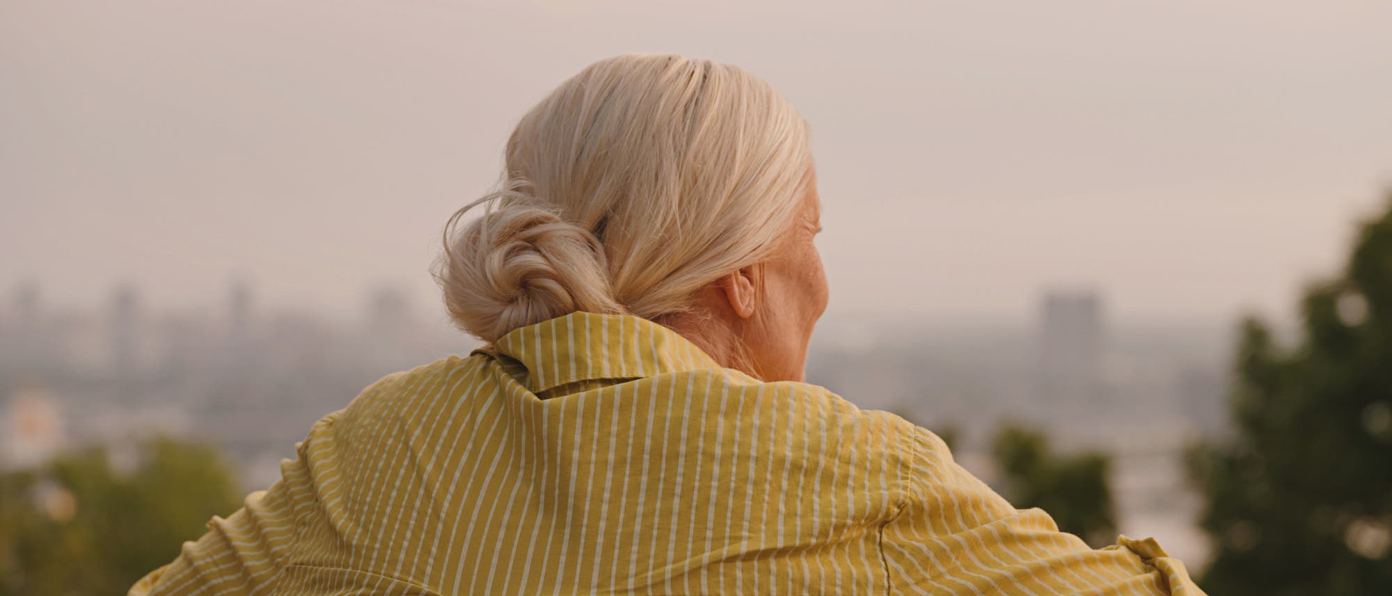 Eine ältere Person mit weißem Haar und einem Dutt steht mit dem Rücken zur Kamera und blickt auf eine verschwommene Stadtlandschaft in der Ferne. Im Hintergrund sind Bäume und Gebäude zu erkennen.