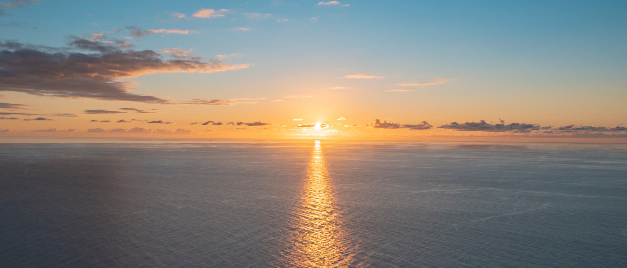 Sonnenuntergang über dem Meer, mit einem klaren Himmel und vereinzelten Wolken. Die Sonne wirft einen goldenen Lichtstreifen auf die Wasseroberfläche, der sich bis zum Horizont erstreckt. Die Farben des Himmels reichen von Blau bis zu warmen Orangetönen. Sonnenuntergang über dem Meer, mit einem klaren Himmel und vereinzelten Wolken. Die Sonne wirft einen goldenen Lichtstreifen auf die Wasseroberfläche, der sich bis zum Horizont erstreckt. Die Farben des Himmels reichen von Blau bis zu warmen Orangetönen.