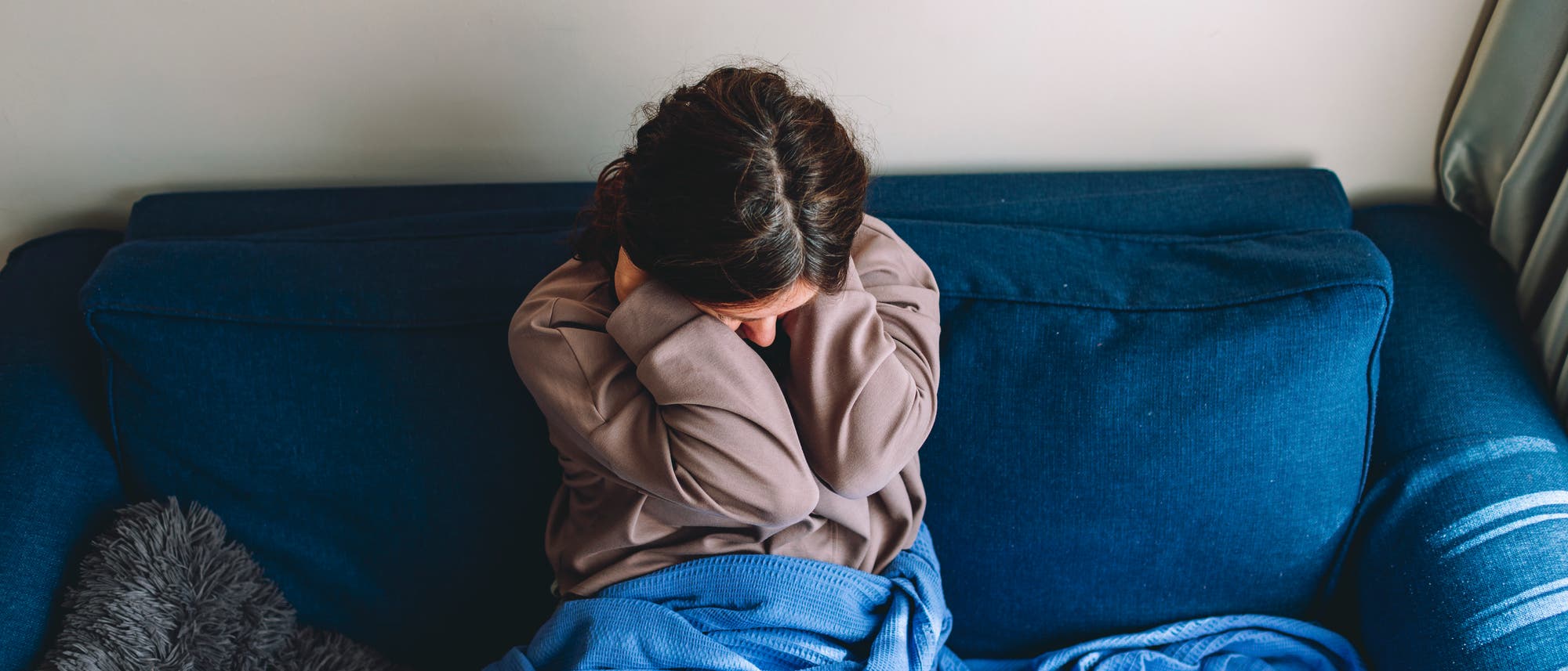 Junge Frau sitzt auf einem blauen Sofa und hält sich den Nacken mit beiden Händen