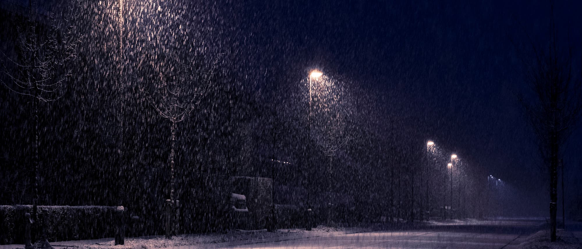Eine n&auml;chtliche Stra&szlig;enszene mit starkem Schneefall. Mehrere Stra&szlig;enlaternen beleuchten die schneebedeckte Stra&szlig;e, w&auml;hrend die Schneeflocken im Licht sichtbar sind. Die Umgebung ist dunkel, und die Stra&szlig;e wirkt verlassen.