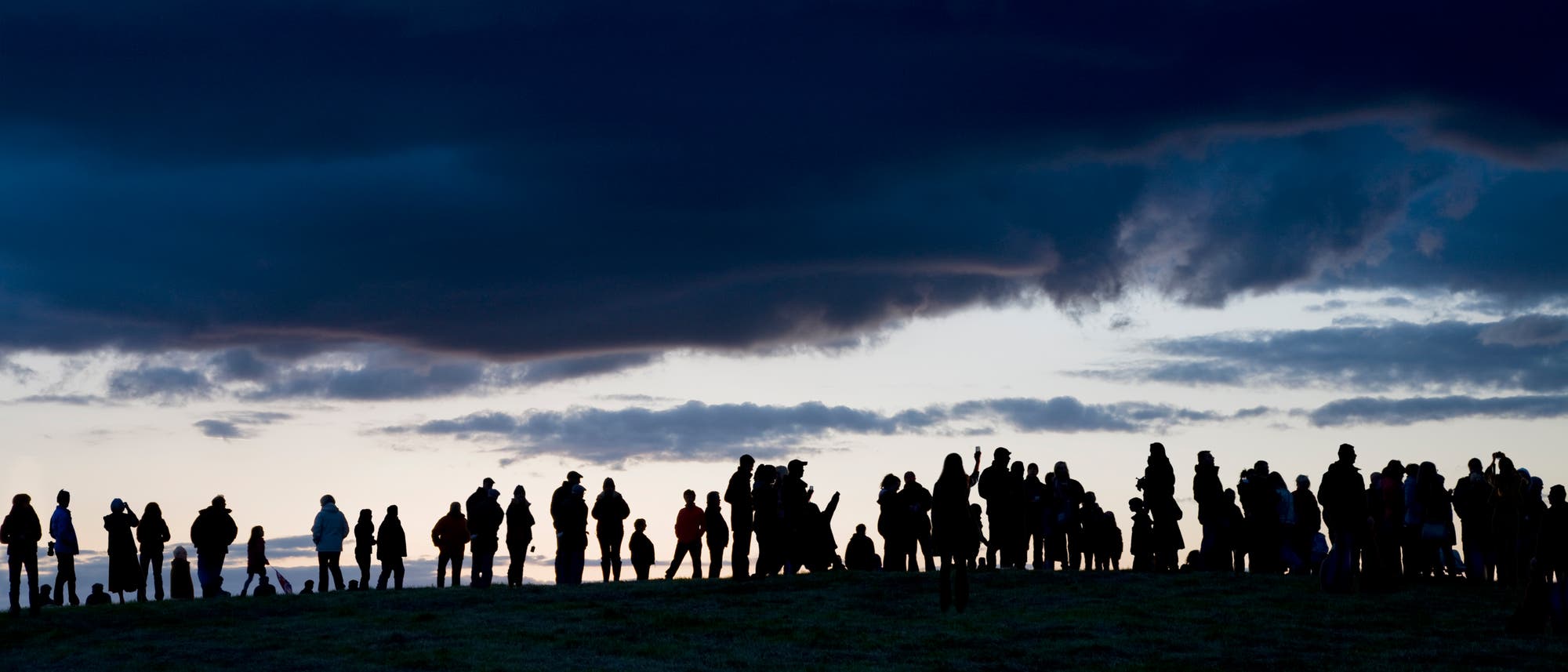Eine Gruppe von Menschen steht auf einem H&uuml;gel und blickt in die Ferne. Der Himmel ist von dunklen Wolken bedeckt, w&auml;hrend der Horizont in der D&auml;mmerung erleuchtet ist. Die Personen sind als Silhouetten vor dem Hintergrund des dramatischen Himmels zu sehen.