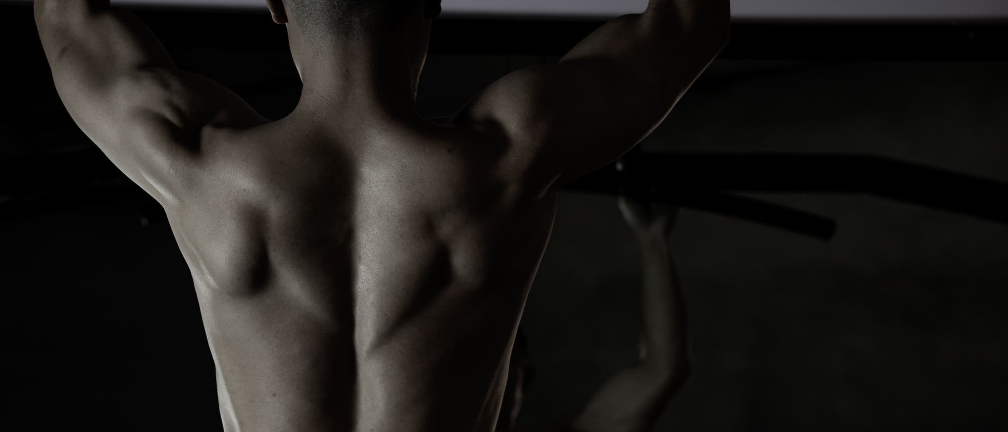 Eine Person mit muskulösem Rücken macht Klimmzüge an einer Stange in einem Fitnessstudio. Die Szene ist dunkel beleuchtet, wodurch die Definition der Rückenmuskulatur hervorgehoben wird. Es sind keine weiteren Details oder Texte im Bild vorhanden. Eine Person mit muskulösem Rücken macht Klimmzüge an einer Stange in einem Fitnessstudio. Die Szene ist dunkel beleuchtet, wodurch die Definition der Rückenmuskulatur hervorgehoben wird. Es sind keine weiteren Details oder Texte im Bild vorhanden.