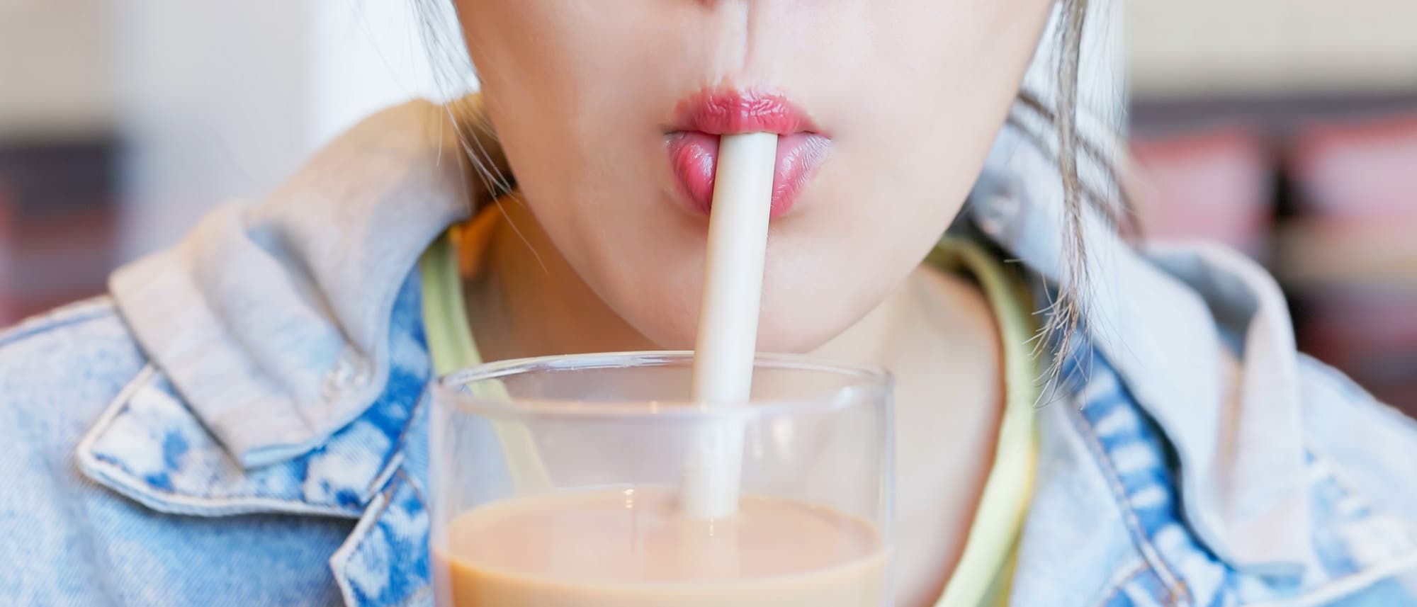 Eine Person in einer Jeansjacke trinkt einen Milchtee mit einem breiten Strohhalm aus einem Glas. Der Fokus liegt auf den Lippen und dem Strohhalm. Im Hintergrund sind verschwommene Details eines Cafés zu erkennen.