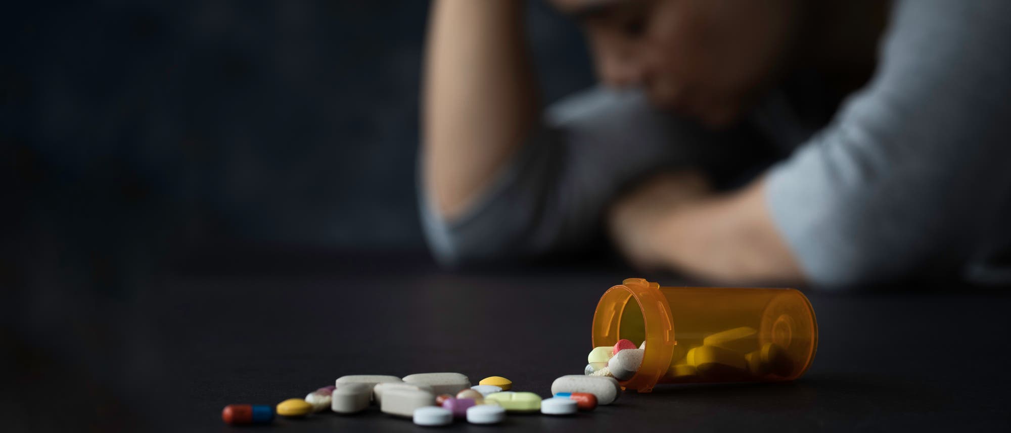 Eine Person sitzt mit gesenktem Kopf im Hintergrund, während im Vordergrund ein umgekippter Pillenbehälter mit verstreuten Tabletten zu sehen ist. Die Szene vermittelt ein Gefühl von Nachdenklichkeit oder Besorgnis.