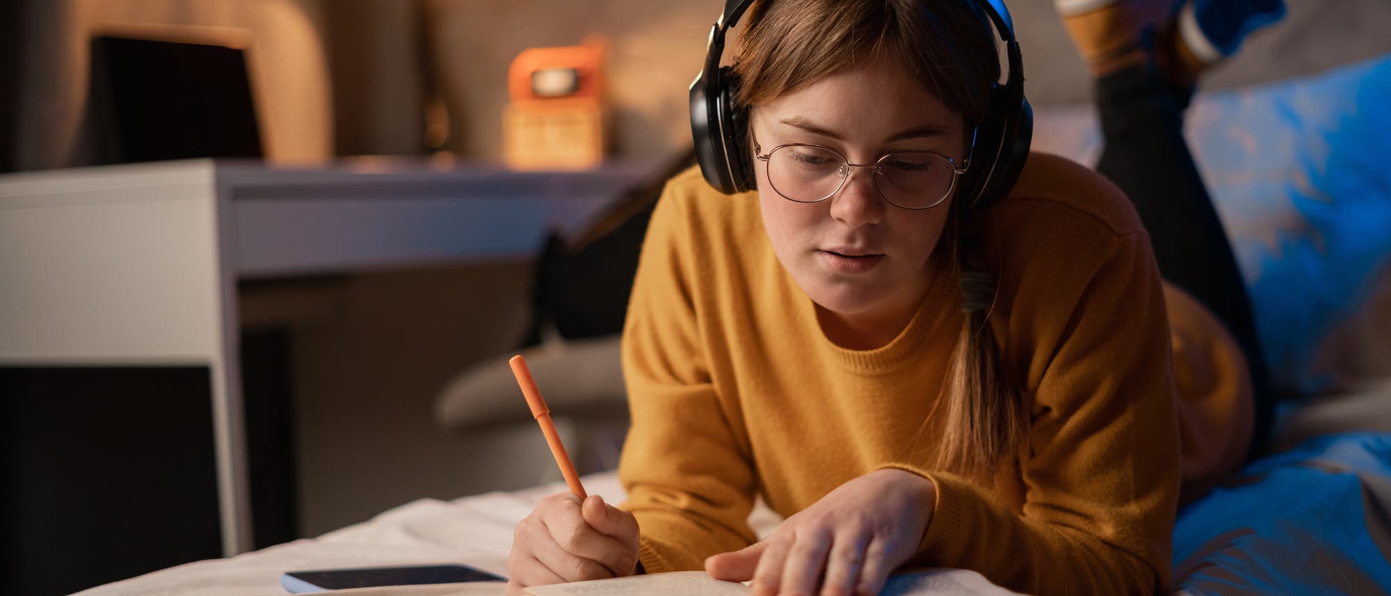 Eine Jugendliche liegt auf einem Bett und trägt Kopfhörer. Sie hält einen Stift in der Hand und schaut konzentriert in ein Buch. Im Hintergrund ist ein Schreibtisch mit einer Lampe und einem Laptop zu sehen. 