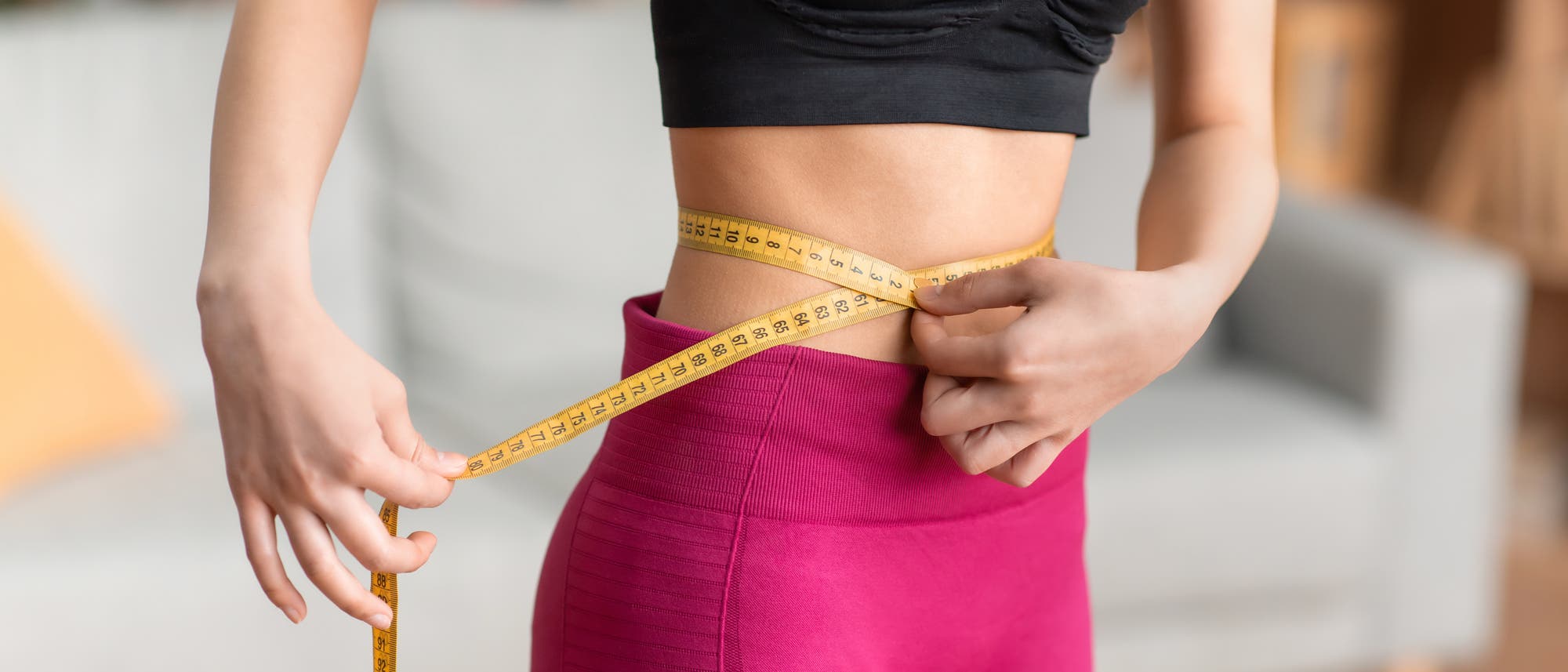 Eine Person in Sportkleidung misst ihren Taillenumfang mit einem gelben Ma&szlig;band. Sie tr&auml;gt ein schwarzes Oberteil und pinke Leggings. Im Hintergrund ist ein unscharfes Wohnzimmer mit einem Sofa zu sehen. Das Bild vermittelt ein Thema von Fitness und K&ouml;rperbewusstsein.