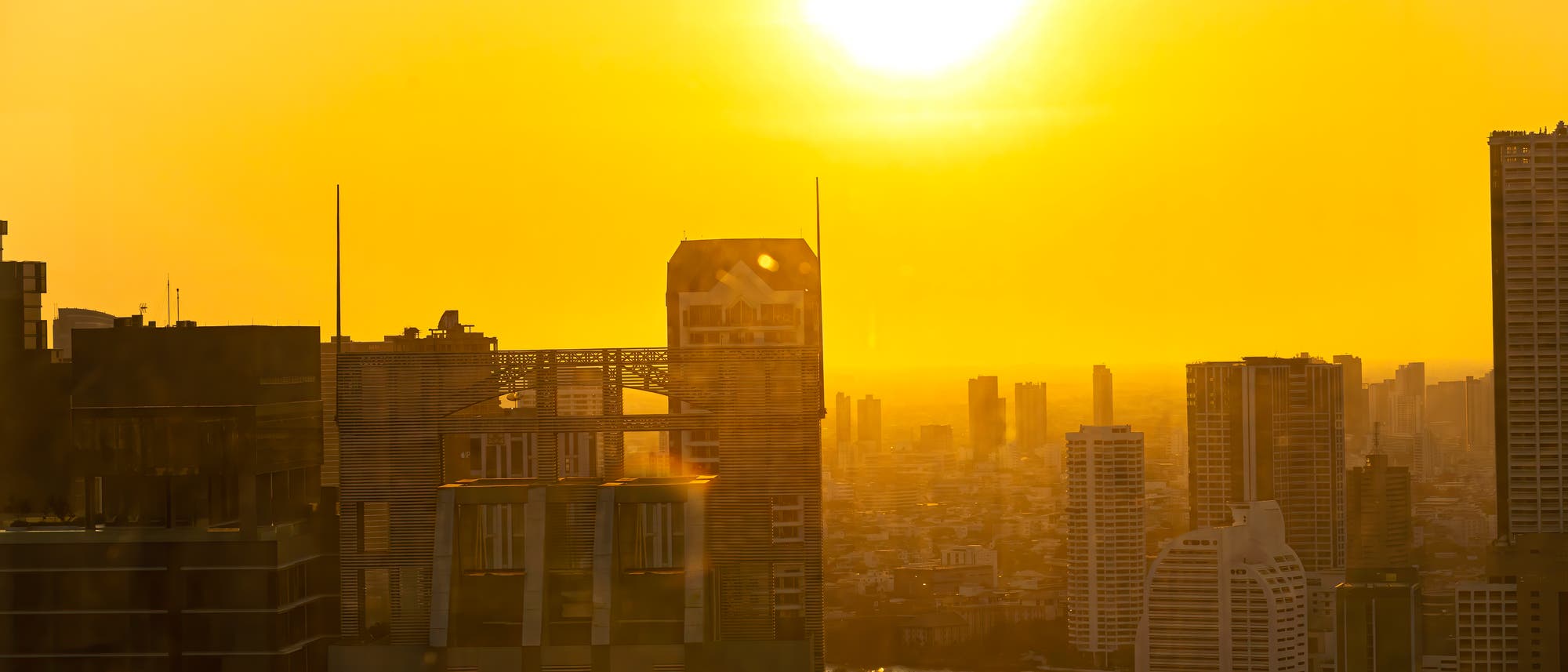 Städtische Skyline bei Sonnenuntergang, mit hohen Gebäuden im Vordergrund und einer leuchtend gelben Sonne am Himmel. Die Szene ist in warmes, goldenes Licht getaucht, das die Silhouetten der Wolkenkratzer betont.