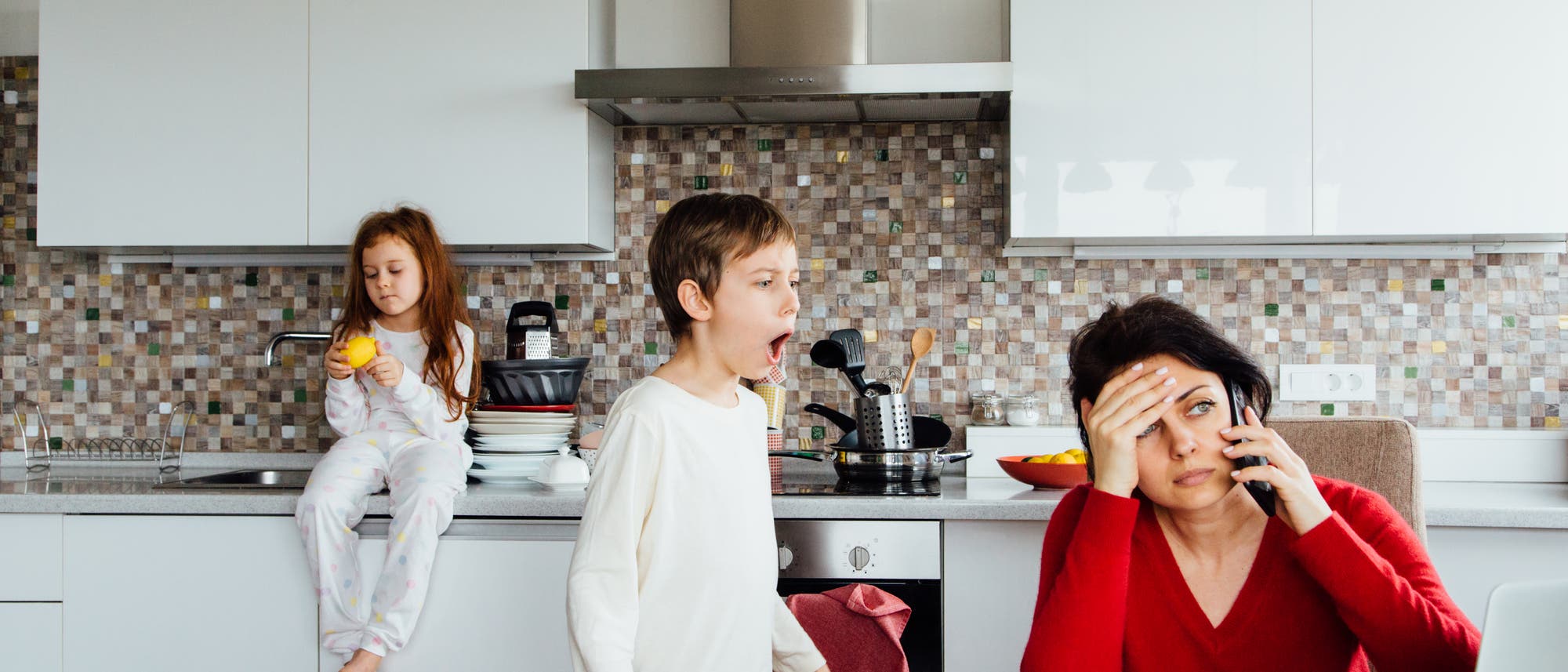 Eine Frau sitzt in einer Küche und telefoniert, während sie sich die Stirn hält, was auf Stress oder Erschöpfung hindeutet. Ein Kind steht neben ihr und scheint zu rufen oder zu singen. Ein weiteres Kind sitzt auf der Arbeitsplatte und hält eine Orange. Die Küche ist modern eingerichtet mit weißen Schränken und einer Mosaikfliesenrückwand. Auf dem Tisch steht eine rote Tasse mit weißen Punkten neben einem Laptop.