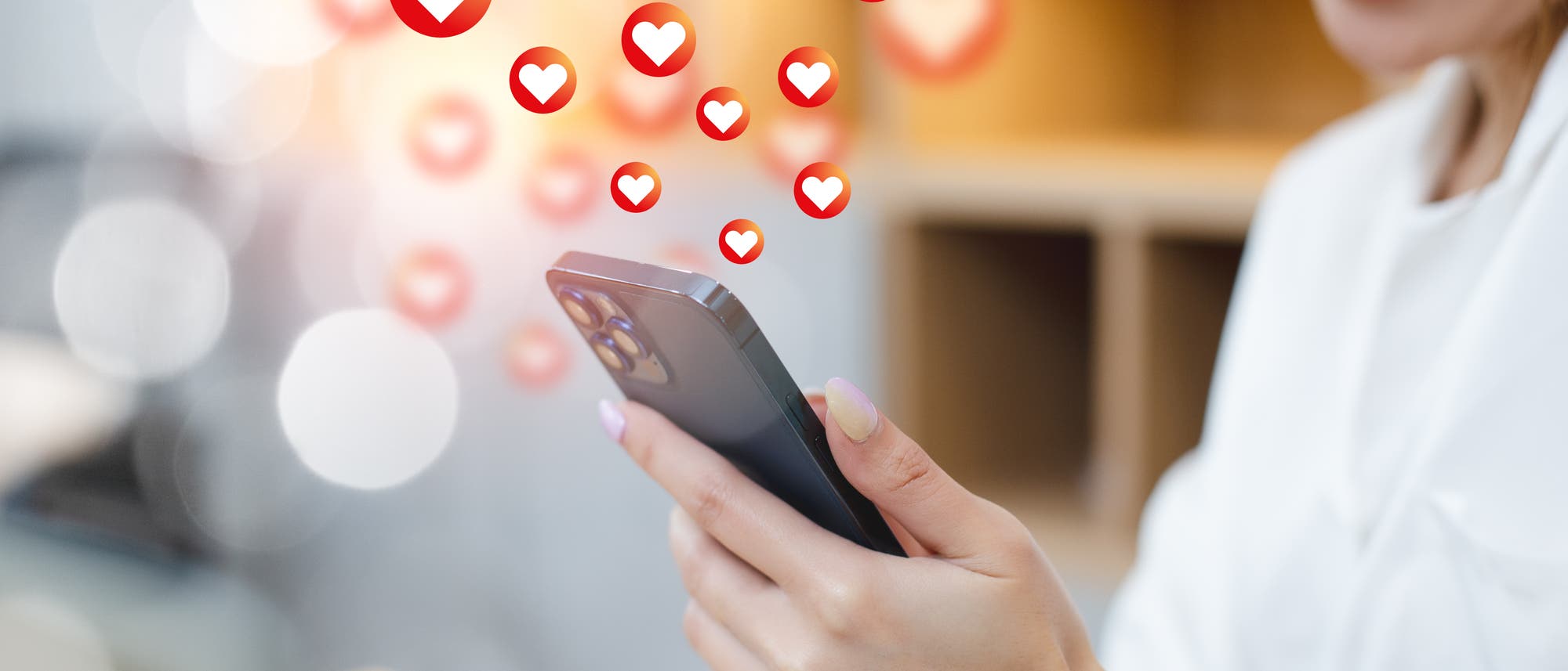 Eine Person h&auml;lt ein Smartphone in der Hand und l&auml;chelt. Auf dem Bildschirm erscheinen mehrere Herzsymbole, die nach oben schweben, was auf soziale Interaktionen oder positive R&uuml;ckmeldungen hinweist. Der Hintergrund ist unscharf, mit warmen Lichtakzenten.