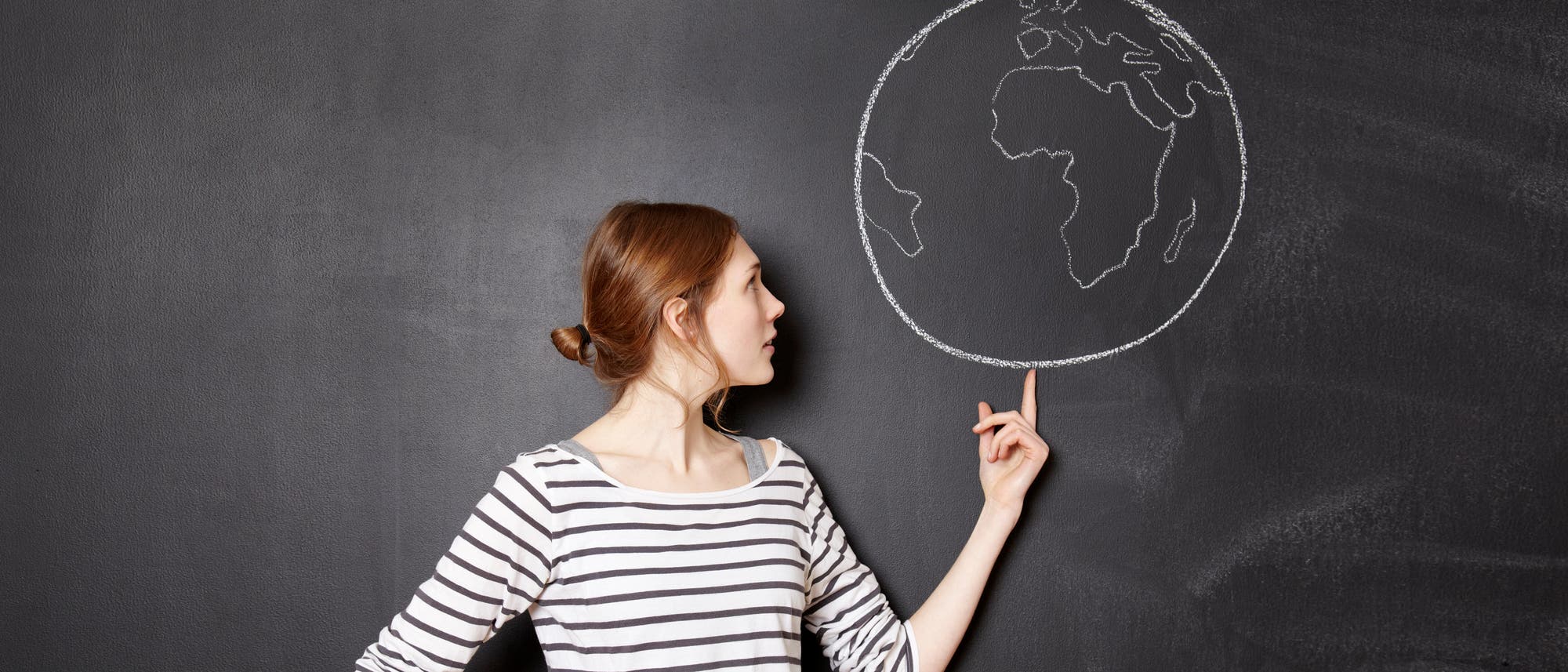 Eine Person in einem gestreiften Oberteil steht vor einer Tafel und zeigt auf eine mit Kreide gezeichnete Skizze der Erde. Die Skizze zeigt die Umrisse von Kontinenten. Die Person blickt zur Zeichnung und scheint in Gedanken vertieft zu sein.
