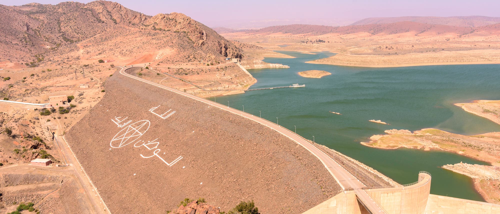 Luftaufnahme eines gro&szlig;en Staudamms in einer trockenen, bergigen Landschaft. Der Damm erstreckt sich &uuml;ber ein Tal und h&auml;lt einen gro&szlig;en, ruhigen Stausee zur&uuml;ck. Auf dem Damm sind in wei&szlig;er Farbe gro&szlig; das Pentagramm aus der Flagge Marokkos und arabische Schriftzeichen geschrieben. Die Umgebung ist karg mit vereinzelten B&uuml;schen und felsigen H&uuml;geln. Der Himmel ist klar und blau.