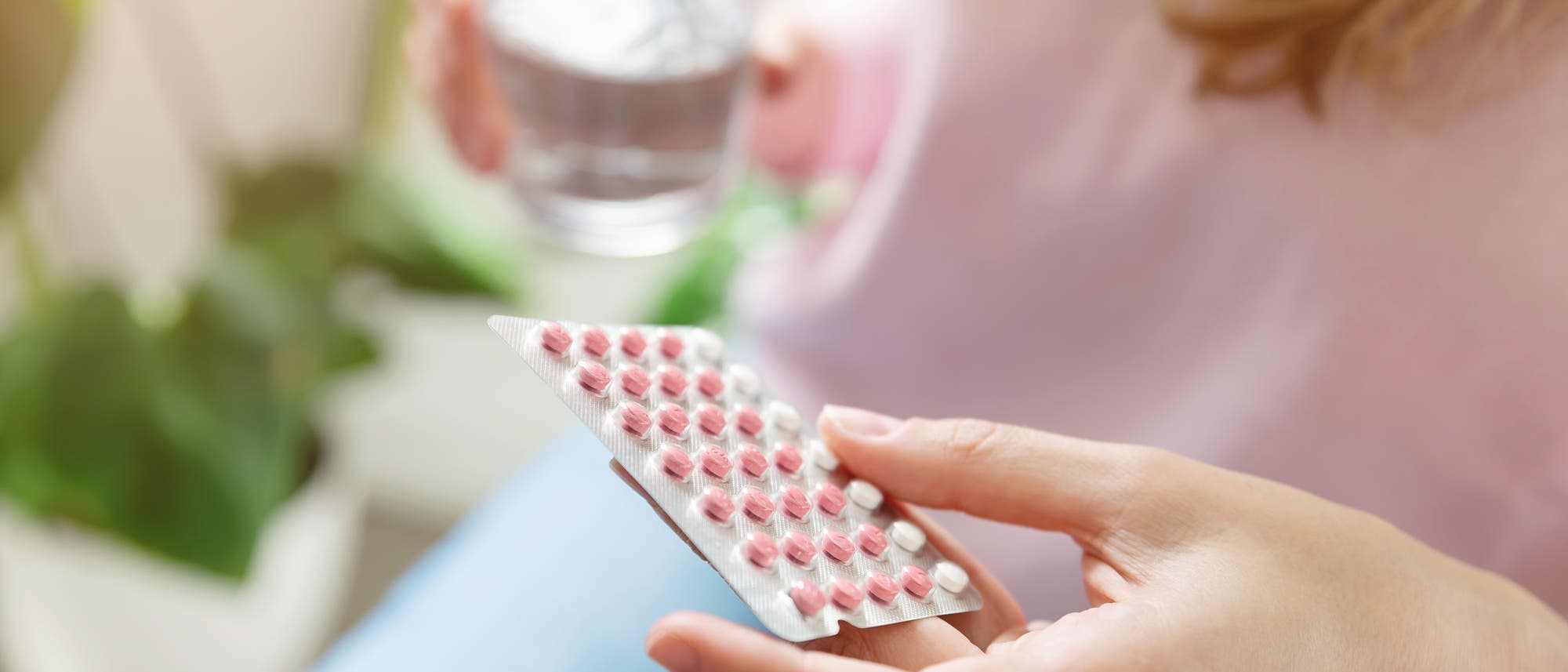 Eine Frau hält eine Blisterpackung mit vier Reihen rosa und einer Reihe weißen Tabletten in der einen Hand und ein Glas Wasser in der anderen. Im Hintergrund sind unscharf grüne Pflanzen zu sehen. Die Szene vermittelt den Eindruck der Einnahme von Medikamenten.