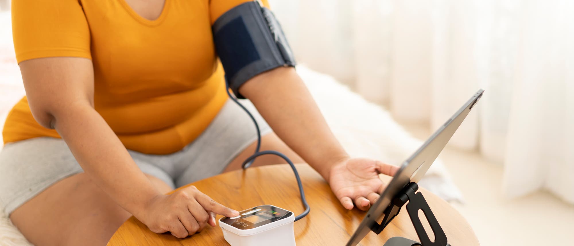 Eine übergewichtige Person in einem orangefarbenen T-Shirt misst ihren Blutdruck mit einem digitalen Blutdruckmessgerät auf einem Holztisch. Der Arm mit der Manschette ruht auf dem Tisch, während die andere Hand das Gerät bedient. Ein Tablet steht auf einem Ständer neben dem Gerät. Die Szene spielt sich in einem hellen Raum mit weißen Vorhängen ab. Eine übergewichtige Person in einem orangefarbenen T-Shirt misst ihren Blutdruck mit einem digitalen Blutdruckmessgerät auf einem Holztisch. Der Arm mit der Manschette ruht auf dem Tisch, während die andere Hand das Gerät bedient. Ein Tablet steht auf einem Ständer neben dem Gerät. Die Szene spielt sich in einem hellen Raum mit weißen Vorhängen ab.