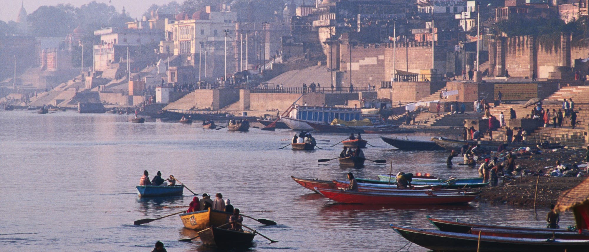 Eine belebte Flusslandschaft mit mehreren Booten, die auf dem Wasser treiben. Im Hintergrund sind historische Gebäude und Treppen zu sehen, die zum Fluss hinabführen. Menschen sind auf den Booten und entlang des Ufers zu erkennen, einige scheinen zu rudern, während andere am Ufer verweilen. Die Szene vermittelt eine geschäftige und kulturell reiche Atmosphäre.