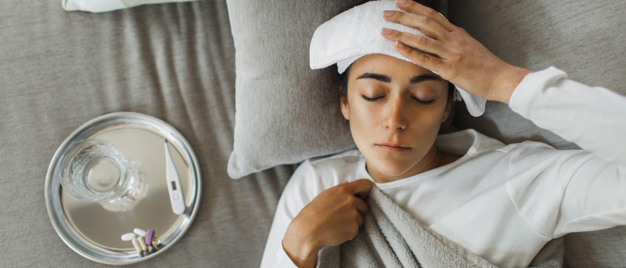 Eine Person liegt auf einem grauen Sofa und hat ein feuchtes Tuch auf der Stirn. Neben ihr steht ein Tablett mit einem Glas Wasser, einem Thermometer und Medikamenten. Die Person ist in eine Decke gehüllt und scheint sich auszuruhen. Die Szene vermittelt den Eindruck von Ruhe und Genesung. Eine Person liegt auf einem grauen Sofa und hat ein feuchtes Tuch auf der Stirn. Neben ihr steht ein Tablett mit einem Glas Wasser, einem Thermometer und Medikamenten. Die Person ist in eine Decke gehüllt und scheint sich auszuruhen. Die Szene vermittelt den Eindruck von Ruhe und Genesung.