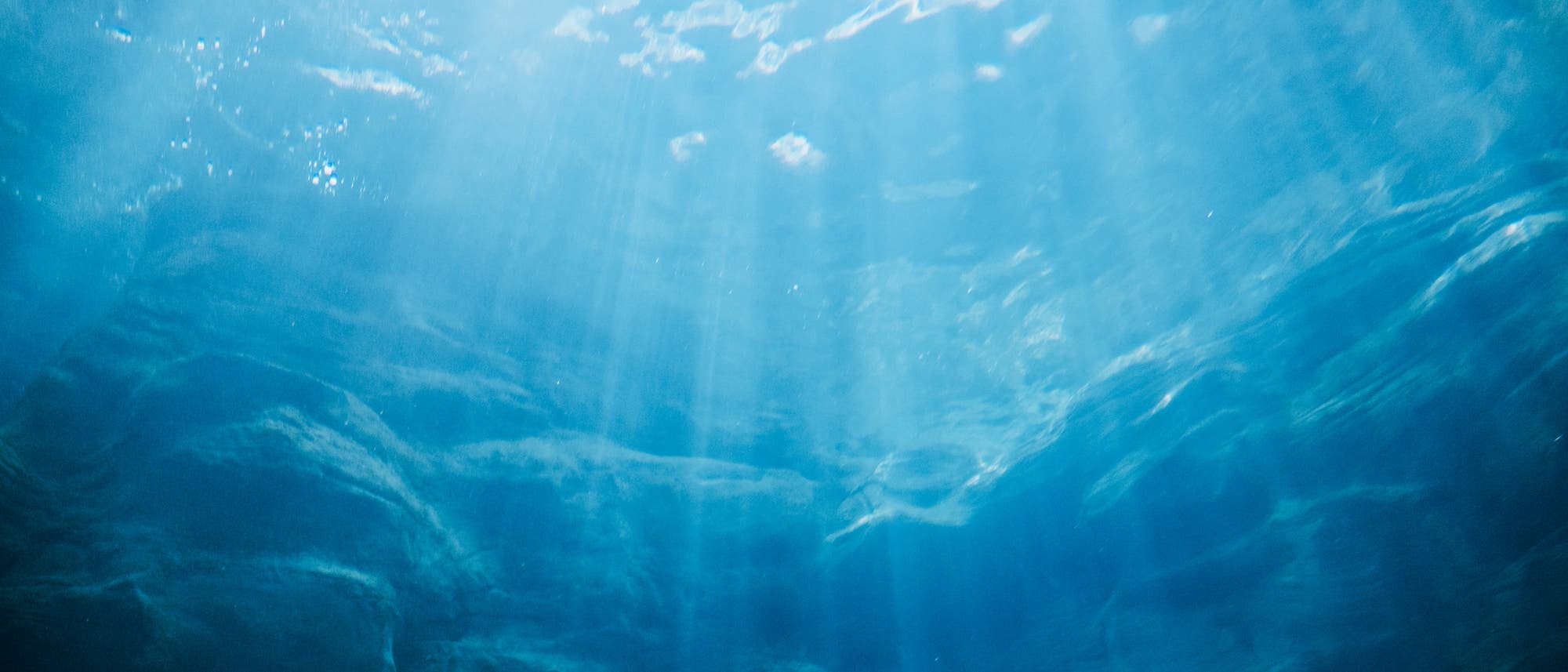 Unterwasseraufnahme eines Ozeans mit Sonnenstrahlen, die durch die Wasseroberfl&auml;che dringen. Die Szene zeigt eine ruhige, blaue Umgebung mit sanften Lichtreflexionen und Schatten auf dem Meeresboden.