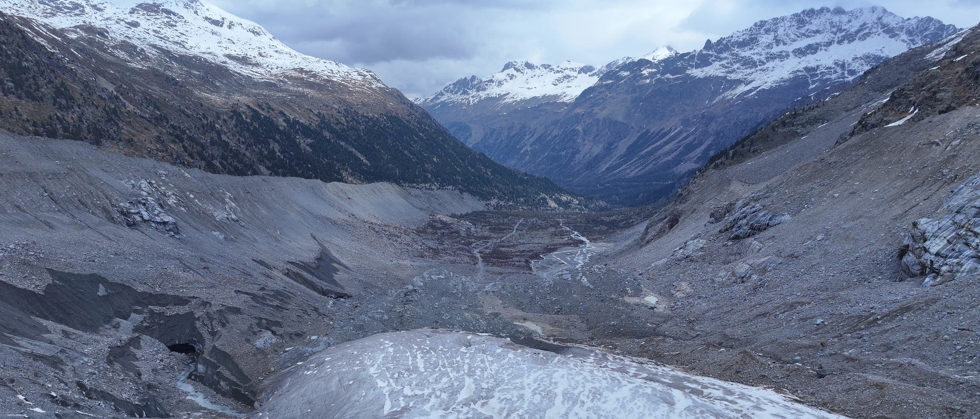 Eine weite Gebirgslandschaft mit schneebedeckten Gipfeln im Hintergrund. Im Vordergrund ist ein Gletscher zu sehen, der sich durch ein felsiges Tal erstreckt. Die Umgebung ist karg und steinig, mit vereinzelten Schneeflecken. Der Himmel ist bewölkt, was auf kühles Wetter hindeutet. Die Szene vermittelt eine ruhige, aber raue alpine Atmosphäre. Eine weite Gebirgslandschaft mit schneebedeckten Gipfeln im Hintergrund. Im Vordergrund ist ein Gletscher zu sehen, der sich durch ein felsiges Tal erstreckt. Die Umgebung ist karg und steinig, mit vereinzelten Schneeflecken. Der Himmel ist bewölkt, was auf kühles Wetter hindeutet. Die Szene vermittelt eine ruhige, aber raue alpine Atmosphäre.