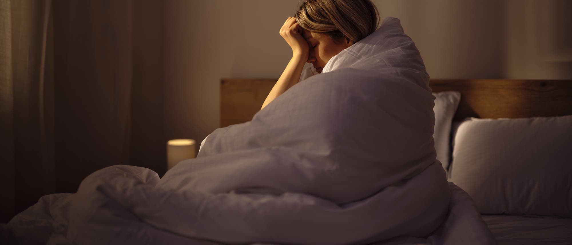 Eine junge Frau sitzt in eine Decke gewickelt im Bett und hält sich den Kopf