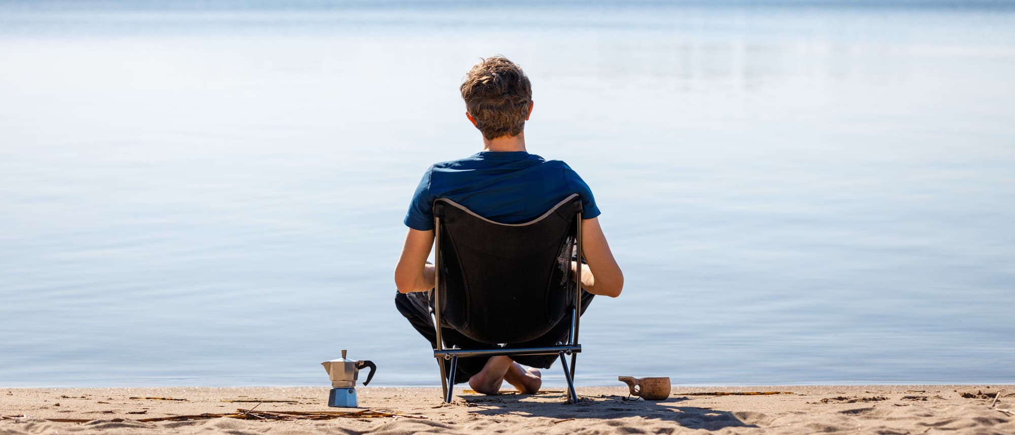 Eine Person sitzt in einem Campingstuhl am Ufer eines ruhigen Sees und blickt auf das Wasser. Neben dem Stuhl stehen eine kleine Kaffeekanne und eine Tasse im Sand. Die Szene vermittelt Ruhe und Entspannung in einer natürlichen Umgebung. Eine Person sitzt in einem Campingstuhl am Ufer eines ruhigen Sees und blickt auf das Wasser. Neben dem Stuhl stehen eine kleine Kaffeekanne und eine Tasse im Sand. Die Szene vermittelt Ruhe und Entspannung in einer natürlichen Umgebung.