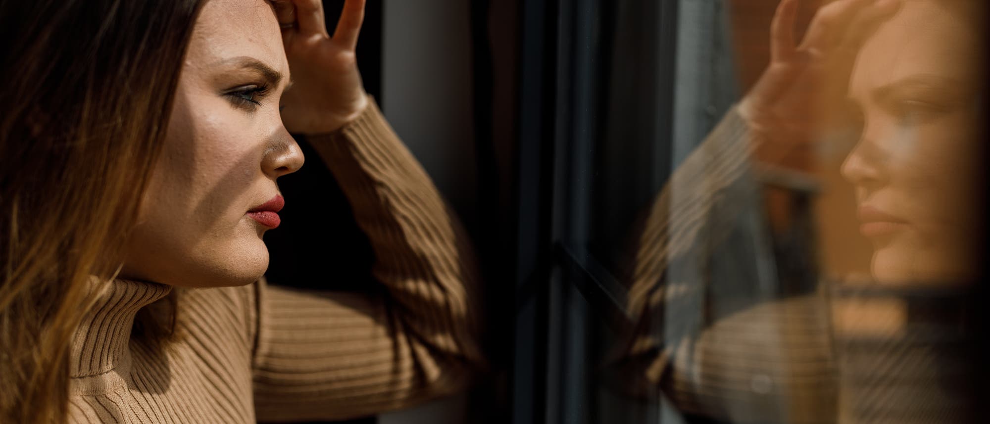 Eine Frau in einem beigefarbenen Rollkragenpullover steht nachdenklich an einem Fenster und schaut hinaus. Ihr Spiegelbild ist im Glas sichtbar. Die Szene ist in warmes Licht getaucht, was eine nachdenkliche und ruhige Atmosphäre schafft. Eine Frau in einem beigefarbenen Rollkragenpullover steht nachdenklich an einem Fenster und schaut hinaus. Ihr Spiegelbild ist im Glas sichtbar. Die Szene ist in warmes Licht getaucht, was eine nachdenkliche und ruhige Atmosphäre schafft.