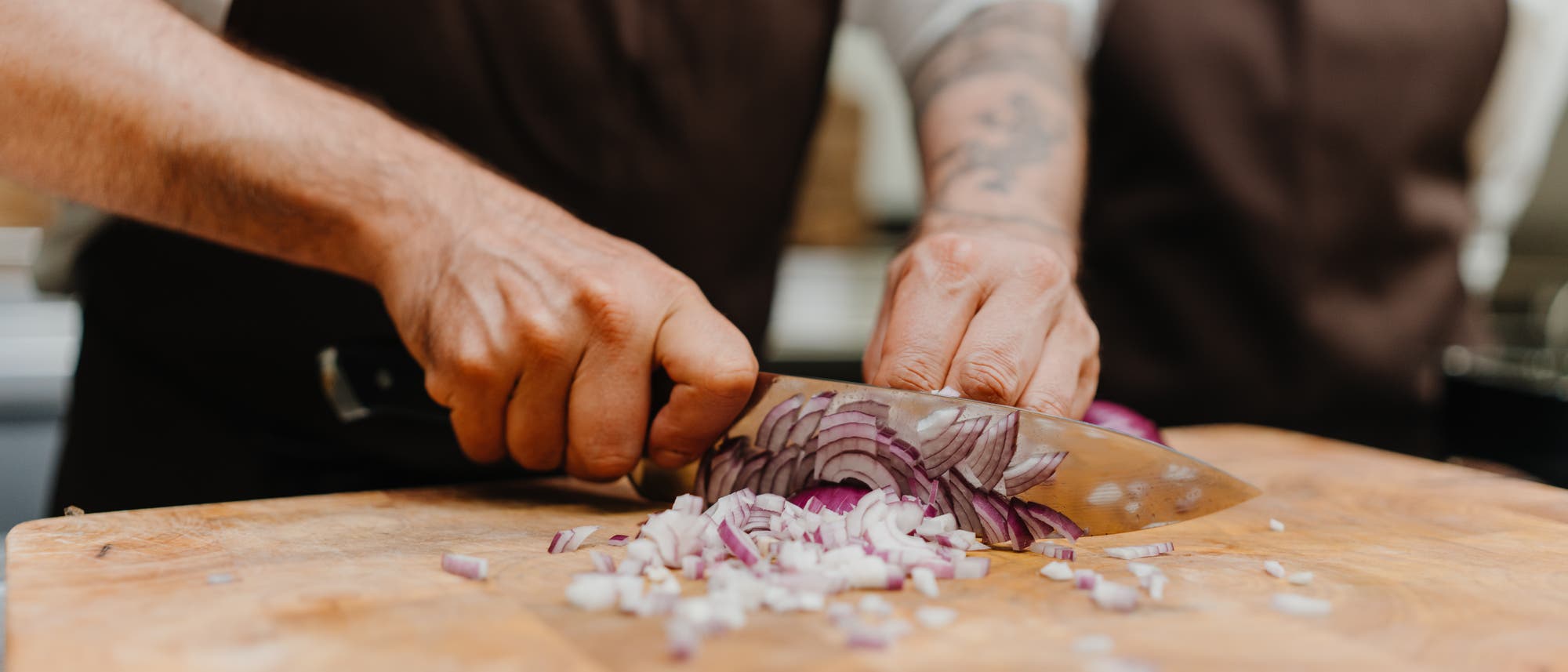 Eine Person schneidet eine rote Zwiebel auf einem Holzbrett mit einem großen Küchenmesser. Die Zwiebel ist in feine Würfel geschnitten. Die Person trägt eine braune Schürze. Im Hintergrund ist eine weitere Person unscharf zu sehen.