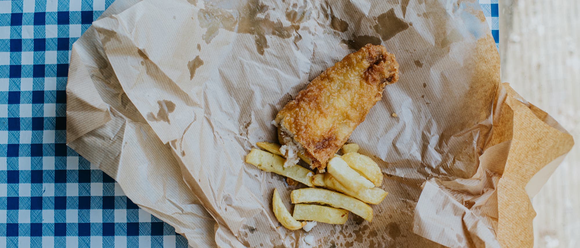 Ein St&uuml;ck gebratener Fisch und einige Pommes Frites liegen auf braunem Papier, das auf einem blau-wei&szlig; karierten Tischdeckchen ausgebreitet ist. Das Papier zeigt Fettflecken, die auf die Frische und Fettigkeit des Essens hinweisen.