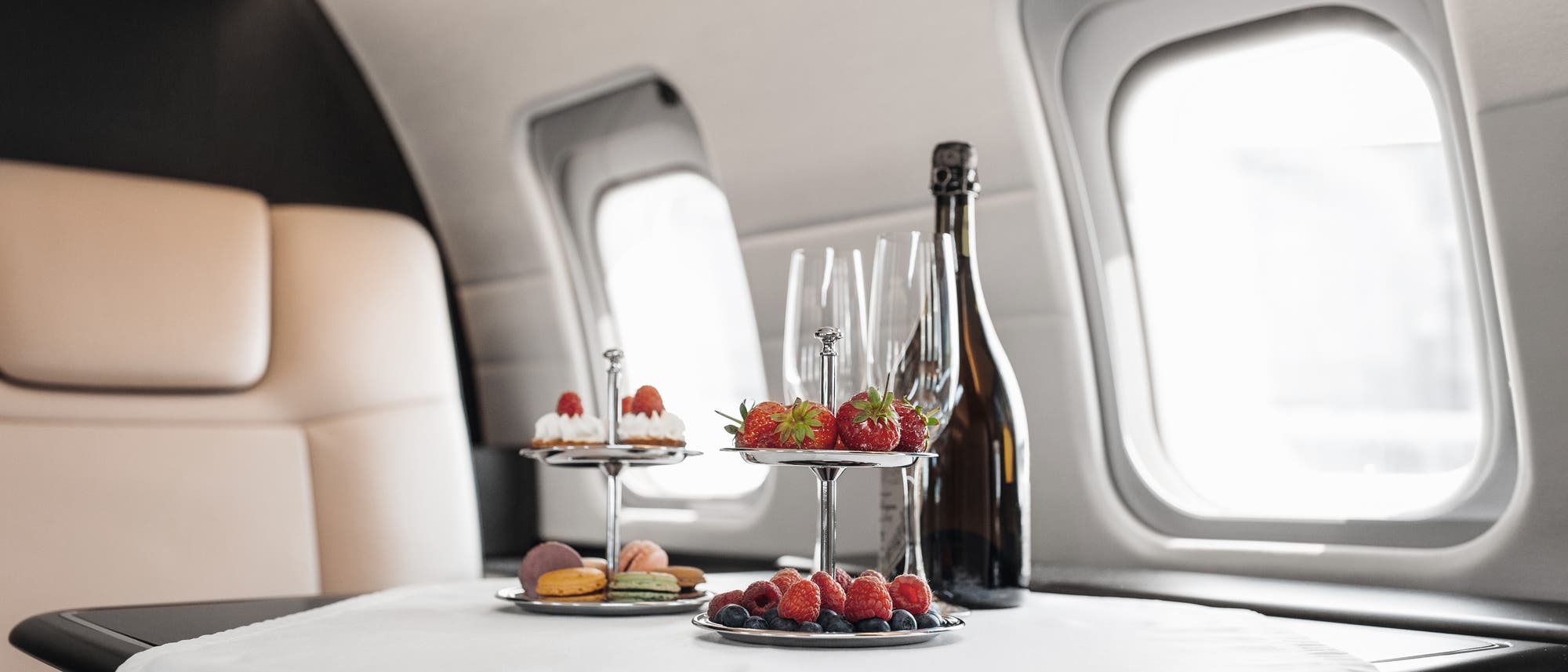 Ein luxuriös gedeckter Tisch in einem Flugzeug mit zwei Etageren, die mit Erdbeeren, Himbeeren und Macarons dekoriert sind. Daneben stehen leere Sektgläser und eine Flasche Champagner. Im Hintergrund sind zwei Flugzeugfenster zu sehen, die Tageslicht hereinlassen. Der Tisch ist mit einem weißen Tuch bedeckt. Ein luxuriös gedeckter Tisch in einem Flugzeug mit zwei Etageren, die mit Erdbeeren, Himbeeren und Macarons dekoriert sind. Daneben stehen leere Sektgläser und eine Flasche Champagner. Im Hintergrund sind zwei Flugzeugfenster zu sehen, die Tageslicht hereinlassen. Der Tisch ist mit einem weißen Tuch bedeckt.