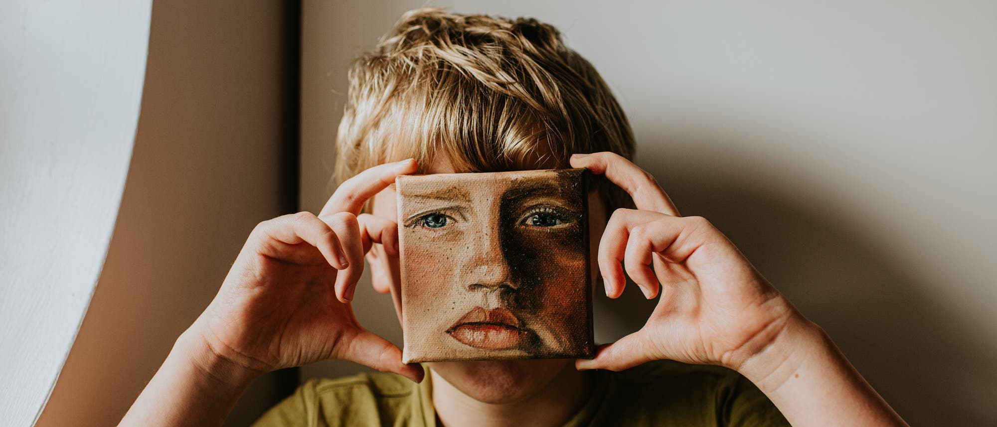 Ein Kind hält ein kleines Gemälde vor sein Gesicht, das ein Porträt zeigt. Das Gemälde verdeckt den oberen Teil des Gesichts des Kindes, sodass die Augen des Porträts anstelle der eigenen Augen erscheinen. Das Kind trägt ein olivgrünes T-Shirt und steht vor einem neutralen Hintergrund. Die Szene spielt mit der Idee von Identität und Kunst. Ein Kind hält ein kleines Gemälde vor sein Gesicht, das ein Porträt zeigt. Das Gemälde verdeckt den oberen Teil des Gesichts des Kindes, sodass die Augen des Porträts anstelle der eigenen Augen erscheinen. Das Kind trägt ein olivgrünes T-Shirt und steht vor einem neutralen Hintergrund. Die Szene spielt mit der Idee von Identität und Kunst.
