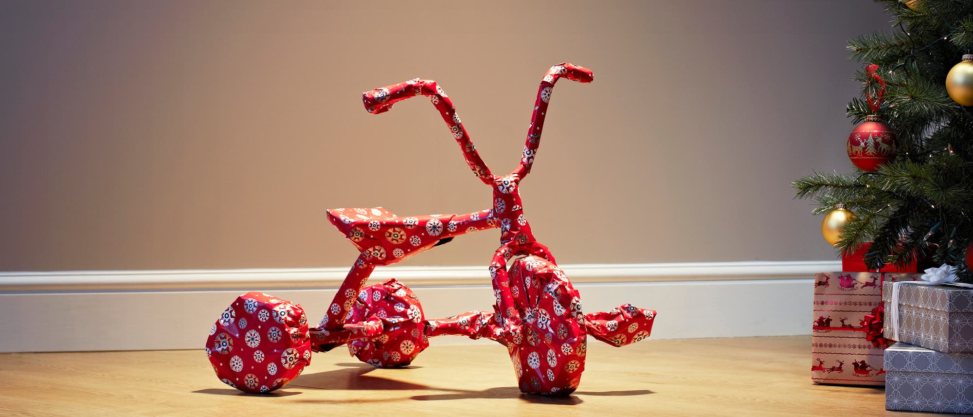 Ein mit rotem Geschenkpapier verpacktes Dreirad steht auf einem Holzboden neben einem geschm&uuml;ckten Weihnachtsbaum. Der Baum ist mit goldenen und roten Kugeln dekoriert, und es liegen mehrere Geschenke darunter. Die Szene vermittelt eine festliche Weihnachtsatmosph&auml;re.