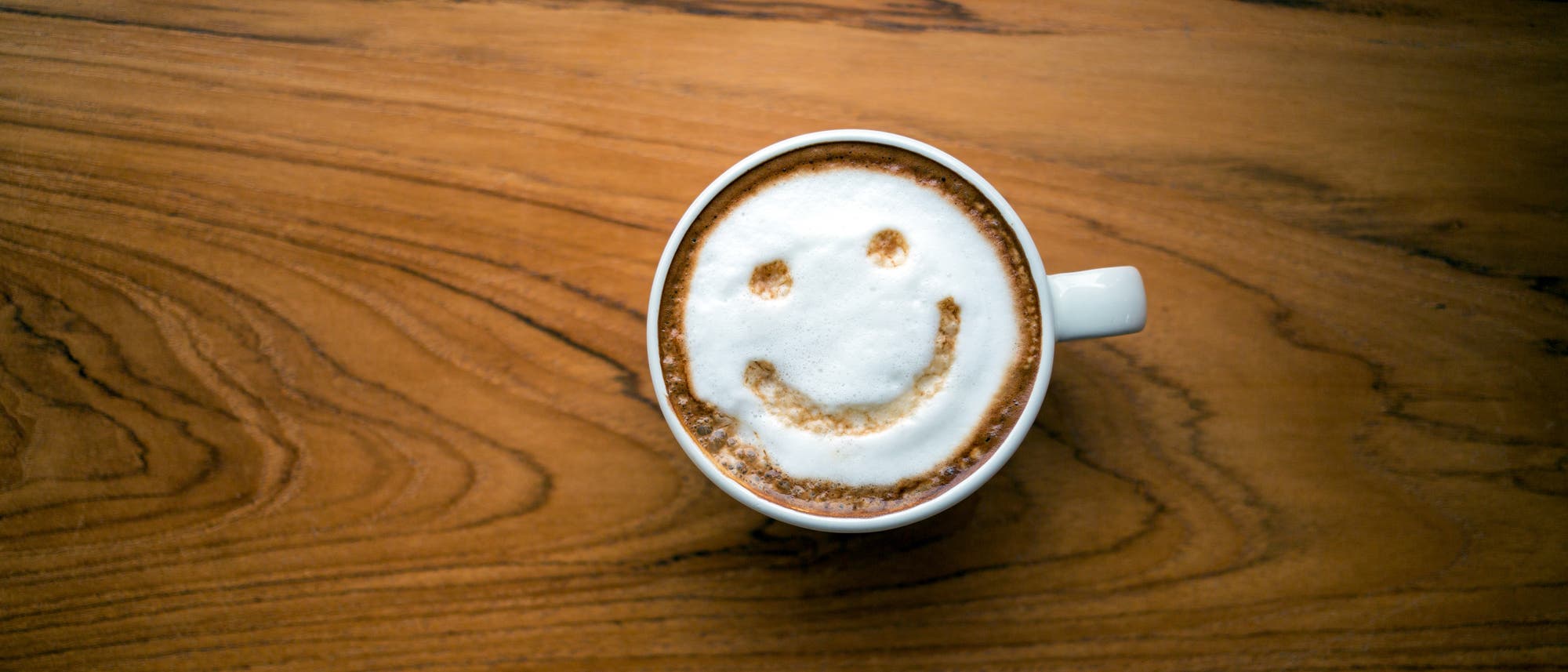 Eine Tasse Cappuccino auf einem Holztisch, mit einem Smiley-Gesicht aus Milchschaum auf der Oberfl&auml;che des Kaffees.