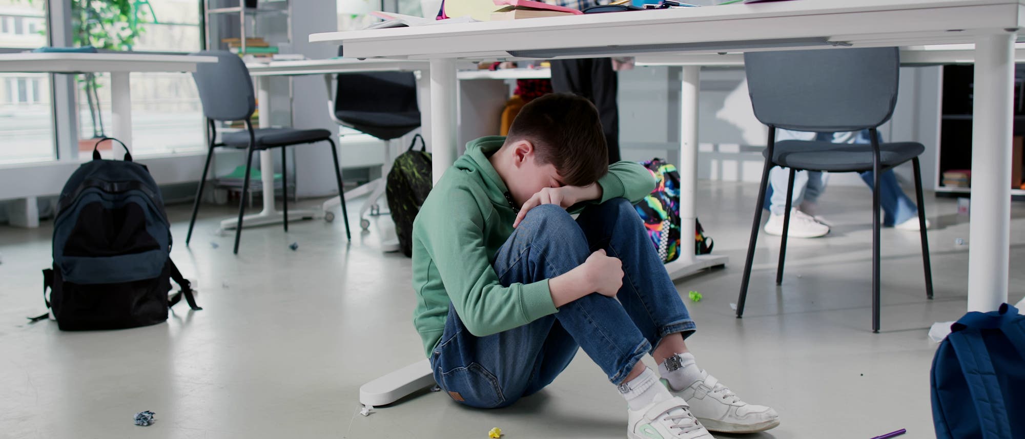 Ein Kind sitzt auf dem Boden eines Klassenzimmers und wirkt traurig. Es hat die Knie angezogen und den Kopf auf die Arme gelegt. Im Hintergrund sind Tische, St&uuml;hle und andere Kinder zu sehen, die besch&auml;ftigt wirken. Auf dem Boden liegen verstreute Papiere und ein Rucksack. Die Szene vermittelt eine Atmosph&auml;re von Isolation und Kummer.