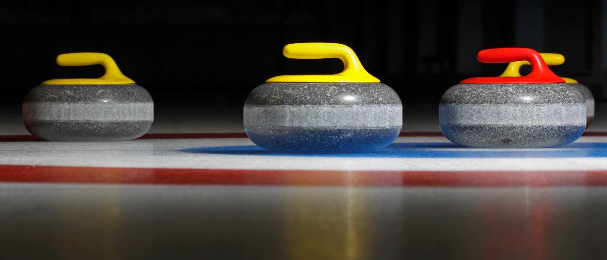 Drei Curling-Steine auf einer Eisbahn, zwei mit gelben Griffen und einer mit einem roten Griff. Die Steine sind auf einem Zielkreis positioniert, der durch rote und blaue Linien markiert ist. Die Szene ist in einer Curling-Halle aufgenommen, die im Hintergrund dunkel ist.