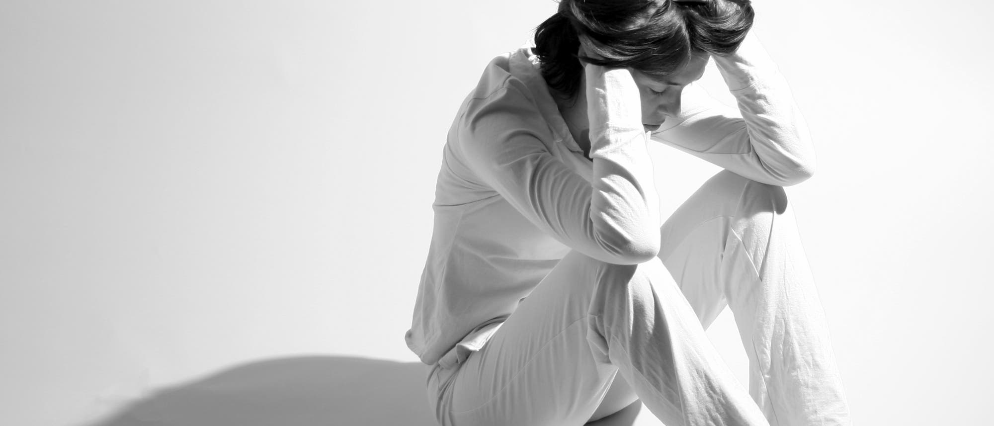 Eine Person sitzt auf dem Boden in einem hellen Raum, die Knie angezogen und den Kopf in den Händen vergraben. Die Person trägt helle Kleidung, und der Schatten fällt deutlich auf den Boden. Die Szene vermittelt einen Ausdruck von Nachdenklichkeit oder Stress. Eine Person sitzt auf dem Boden in einem hellen Raum, die Knie angezogen und den Kopf in den Händen vergraben. Die Person trägt helle Kleidung, und der Schatten fällt deutlich auf den Boden. Die Szene vermittelt einen Ausdruck von Nachdenklichkeit oder Stress.