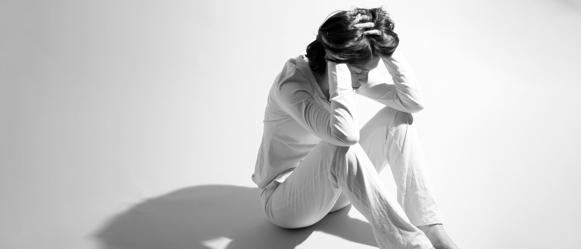Eine Person sitzt auf dem Boden in einem hellen Raum, die Knie angezogen und den Kopf in den Händen vergraben. Die Person trägt helle Kleidung, und der Schatten fällt deutlich auf den Boden. Die Szene vermittelt einen Ausdruck von Nachdenklichkeit oder Stress.
