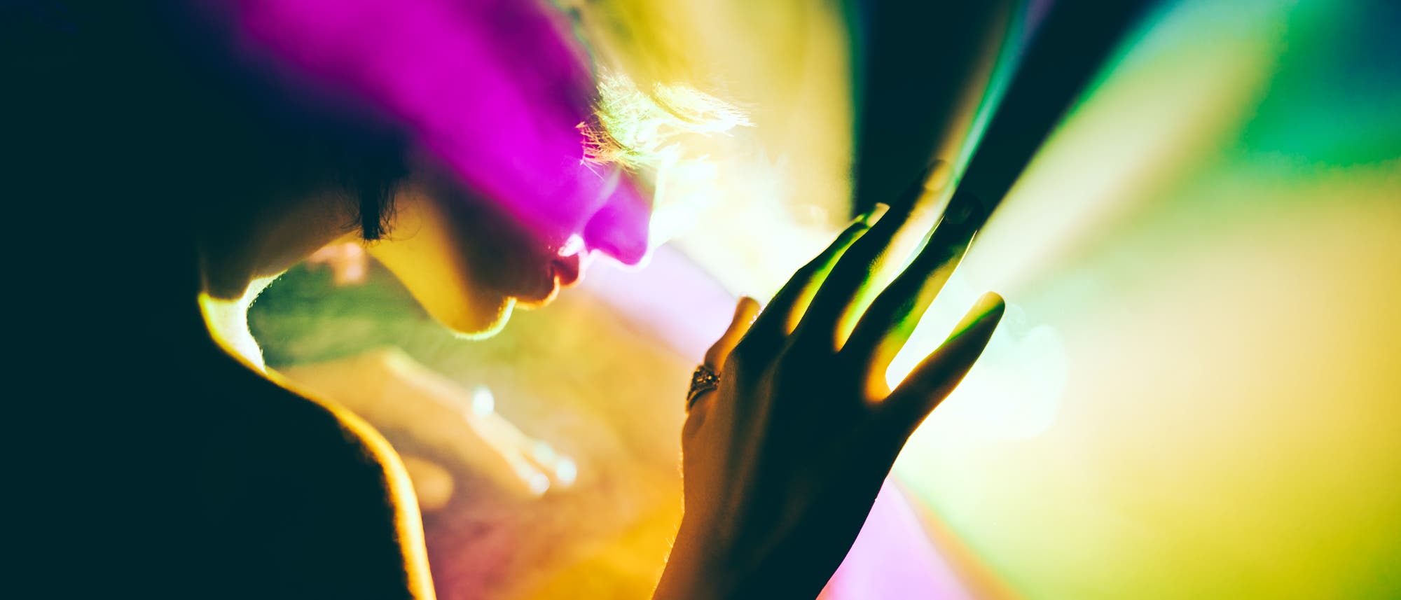 Eine Person in einem künstlerischen Foto, das von bunten Lichtern in Gelb, Grün und Violett beleuchtet wird. Die Silhouette der Person ist seitlich zu sehen, mit einer Hand, die sanft in Richtung der Lichtstrahlen ausgestreckt ist. Die Szene vermittelt eine mystische und träumerische Atmosphäre. Eine Person in einem künstlerischen Foto, das von bunten Lichtern in Gelb, Grün und Violett beleuchtet wird. Die Silhouette der Person ist seitlich zu sehen, mit einer Hand, die sanft in Richtung der Lichtstrahlen ausgestreckt ist. Die Szene vermittelt eine mystische und träumerische Atmosphäre.