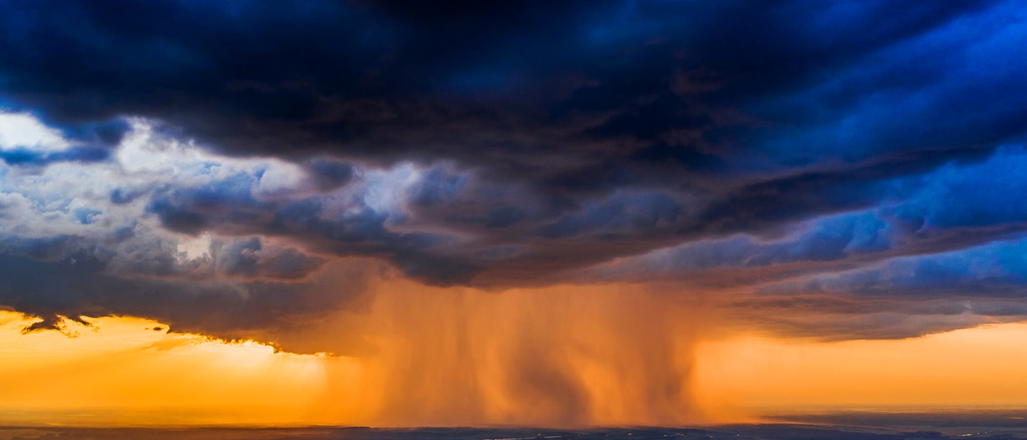 Luftaufnahme eines dramatischen Sonnenuntergangs mit dunklen, bedrohlichen Wolken, die &uuml;ber einer Landschaft h&auml;ngen. Ein intensiver Regenschauer f&auml;llt in der Ferne, w&auml;hrend die untergehende Sonne den Himmel in leuchtenden Orange- und Blaut&ouml;nen f&auml;rbt. Unten sind Fl&uuml;sse und eine Stadtlandschaft zu sehen, die im sanften Licht des Abends schimmern.