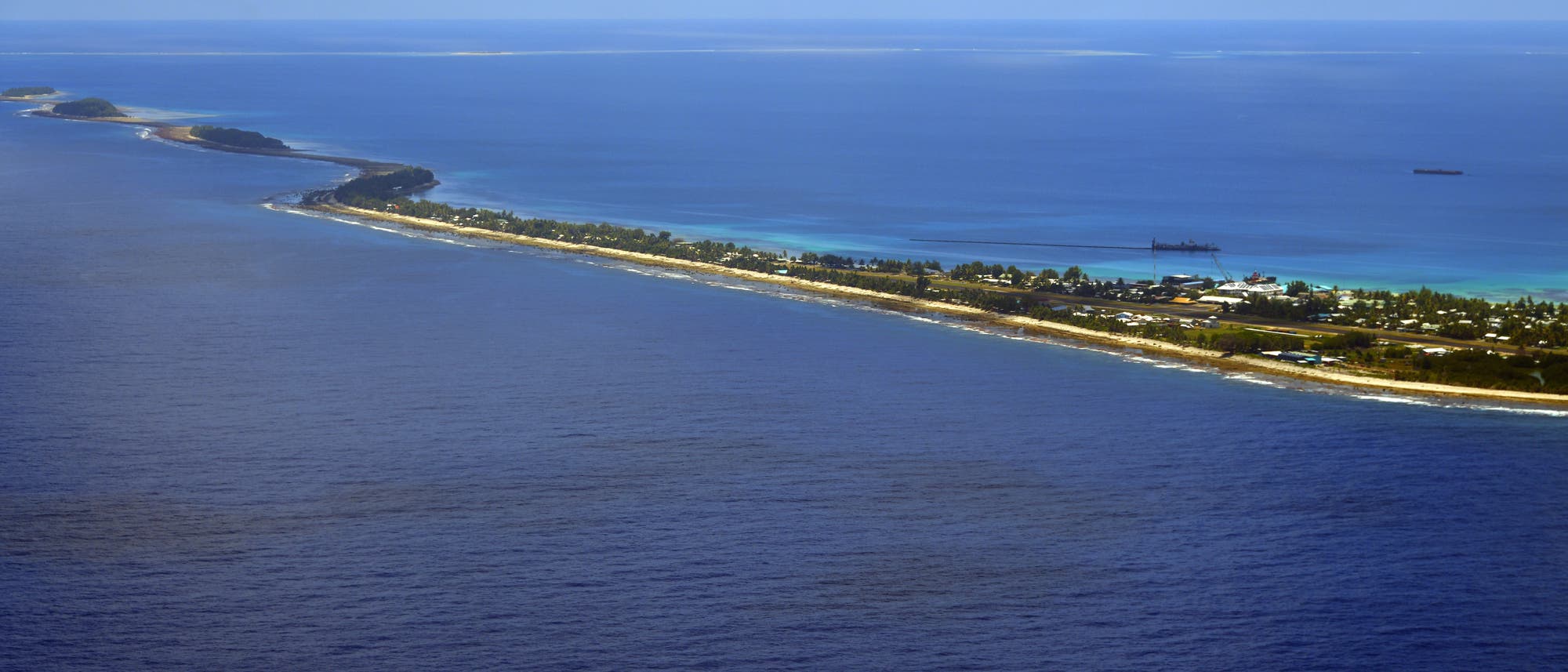 Luftaufnahme einer schmalen, langgestreckten Insel von Tuvalu , umgeben von t&uuml;rkisfarbenem Wasser. Die Insel ist mit gr&uuml;ner Vegetation bedeckt und zeigt eine Ansammlung von Geb&auml;uden entlang der K&uuml;ste. Im Hintergrund sind Wolken am blauen Himmel zu sehen.