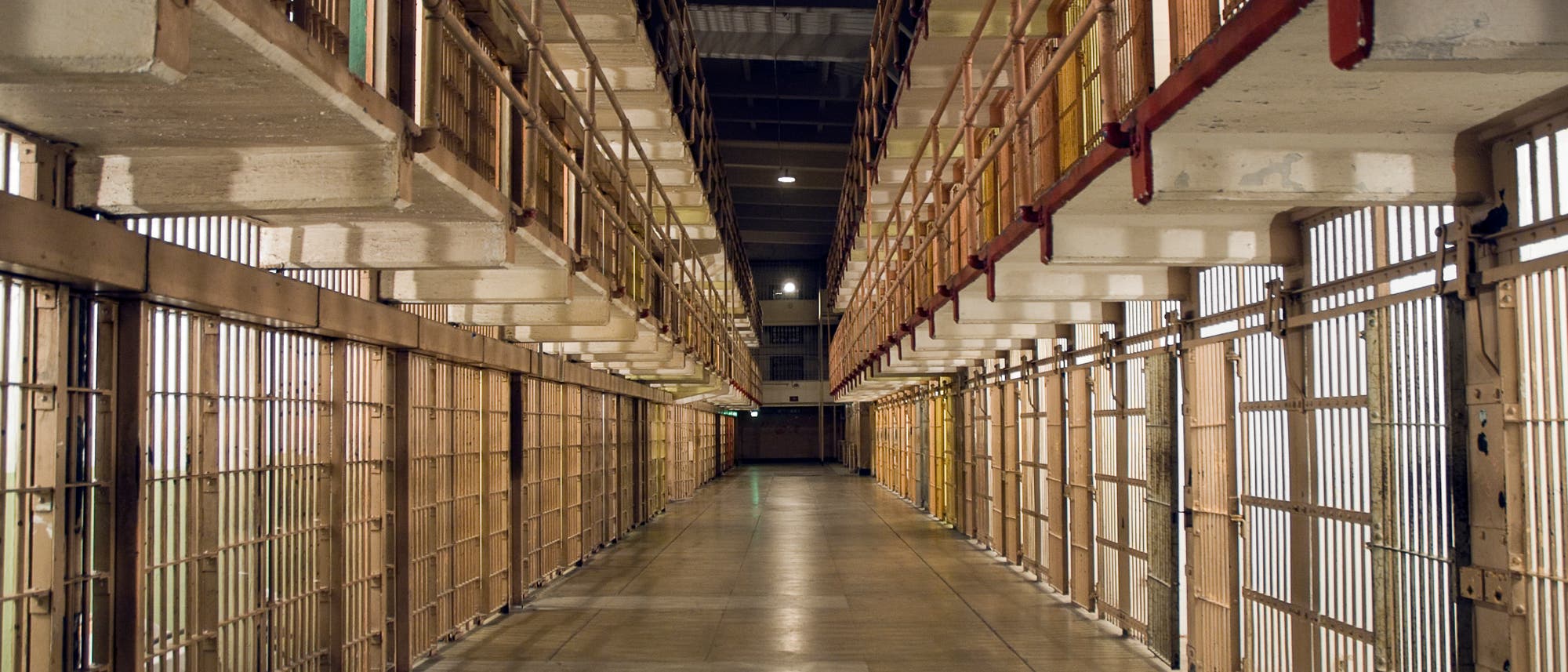 Ein langer, leerer Gefängnisflur mit Zellenreihen auf beiden Seiten. Die Zellen sind durch Gitterstäbe verschlossen, und das Licht fällt durch die Gitter, wodurch ein Muster auf dem Boden entsteht. Die Perspektive des Fotos betont die Tiefe des Flurs.
