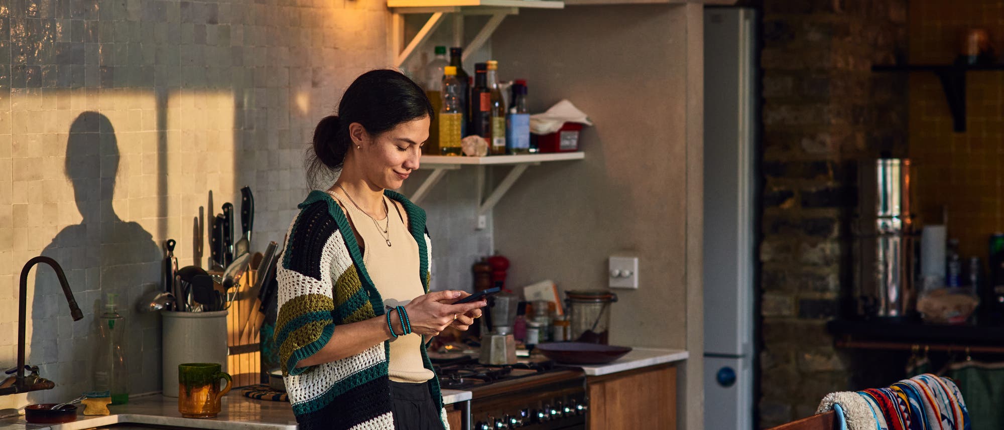 Eine Person steht in einer gemütlichen Küche und schaut auf ihr Smartphone. Die Küche ist mit einem Herd, Regalen mit Gewürzen und einer Spüle ausgestattet. Sonnenlicht fällt durch ein Fenster und wirft Schatten an die Wand. Die Atmosphäre wirkt warm und einladend.