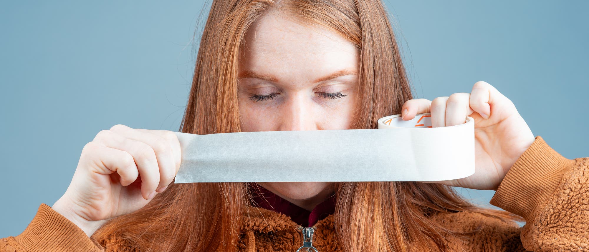 Rothaarige junge Frau klebt sich den Mund mit Panzertape zu.