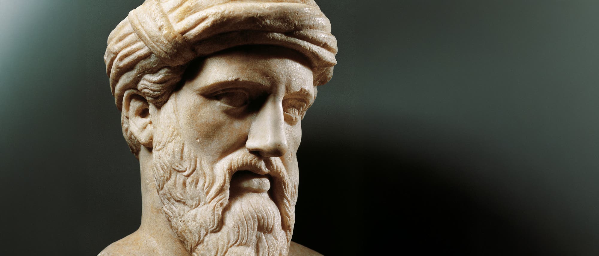 Eine Marmorbüste eines antiken Philosophen mit einem detailliert geschnitzten Bart und einem turbanähnlichen Kopfschmuck. Die Skulptur zeigt einen ernsten Ausdruck und ist vor einem neutralen Hintergrund platziert.