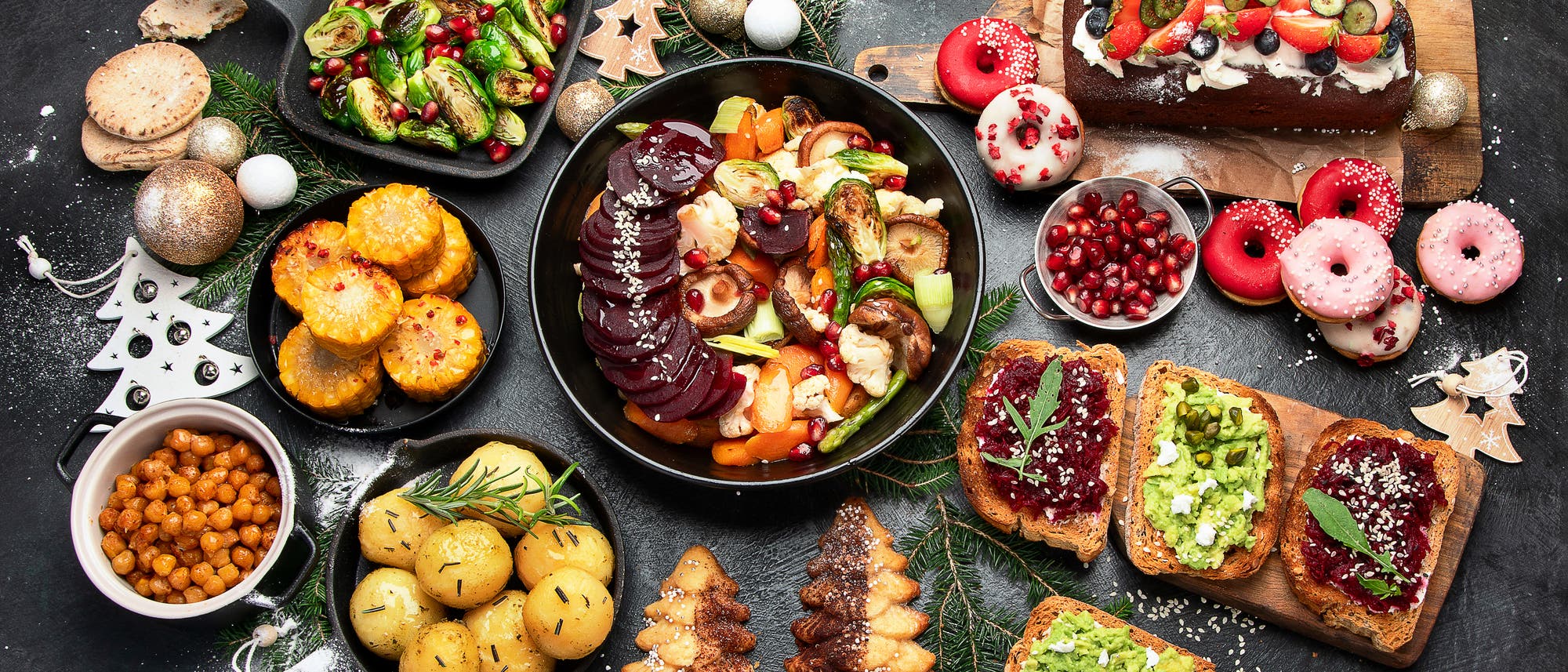 Ein festlich gedeckter Tisch mit einer Vielzahl von Speisen. In der Mitte befindet sich eine Schale mit ger&ouml;stetem Gem&uuml;se, darunter Rote Bete, Karotten und Rosenkohl, garniert mit Granatapfelkernen. Links sind gebratene Kartoffeln und Kichererbsen zu sehen. Rechts liegt ein Holzbrett mit belegten Brotscheiben, dekoriert mit Avocado und Marmelade. Im Hintergrund sind weihnachtliche Dekorationen wie Sterne und Tannenzweige verteilt. Ein Schokoladenkuchen mit frischen Beeren und Donuts erg&auml;nzt das Arrangement.