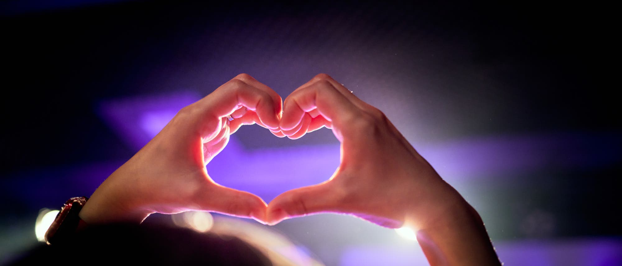 Hände formen ein Herzsymbol vor einem verschwommenen Hintergrund mit lila und blauen Lichtern. Die Szene vermittelt eine positive und liebevolle Stimmung. Hände formen ein Herzsymbol vor einem verschwommenen Hintergrund mit lila und blauen Lichtern. Die Szene vermittelt eine positive und liebevolle Stimmung.