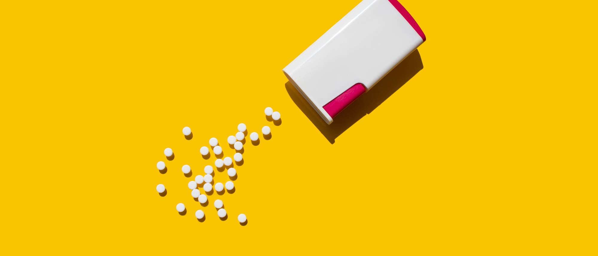Ein wei&szlig;er Beh&auml;lter mit rotem Deckel liegt auf einem gelben Hintergrund. Aus dem Beh&auml;lter sind mehrere kleine, wei&szlig;e Tabletten herausgefallen und verteilen sich auf der Fl&auml;che.