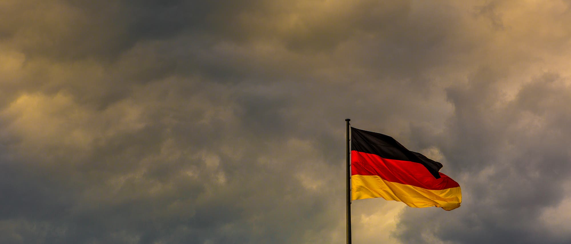 Eine deutsche Flagge weht im Wind vor einem dramatischen, wolkenverhangenen Himmel. Die Farben Schwarz, Rot und Gold sind deutlich sichtbar. Die Szene vermittelt eine Stimmung von Stärke und Beständigkeit. Eine deutsche Flagge weht im Wind vor einem dramatischen, wolkenverhangenen Himmel. Die Farben Schwarz, Rot und Gold sind deutlich sichtbar. Die Szene vermittelt eine Stimmung von Stärke und Beständigkeit.
