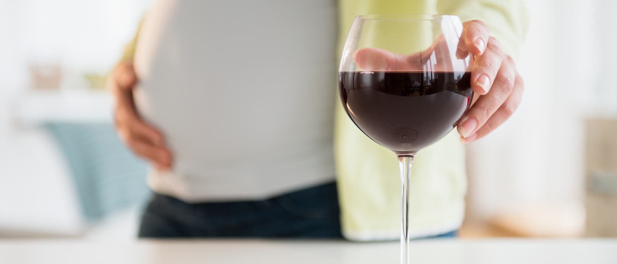 Eine schwangere Person steht an einem Tisch und h&auml;lt ein Glas Rotwein. Die Hand ruht auf dem Glas, w&auml;hrend die andere Hand den Bauch ber&uuml;hrt. Die Szene vermittelt eine nachdenkliche Stimmung in Bezug auf Alkoholkonsum w&auml;hrend der Schwangerschaft. Im Hintergrund ist ein unscharfer Wohnraum zu sehen.