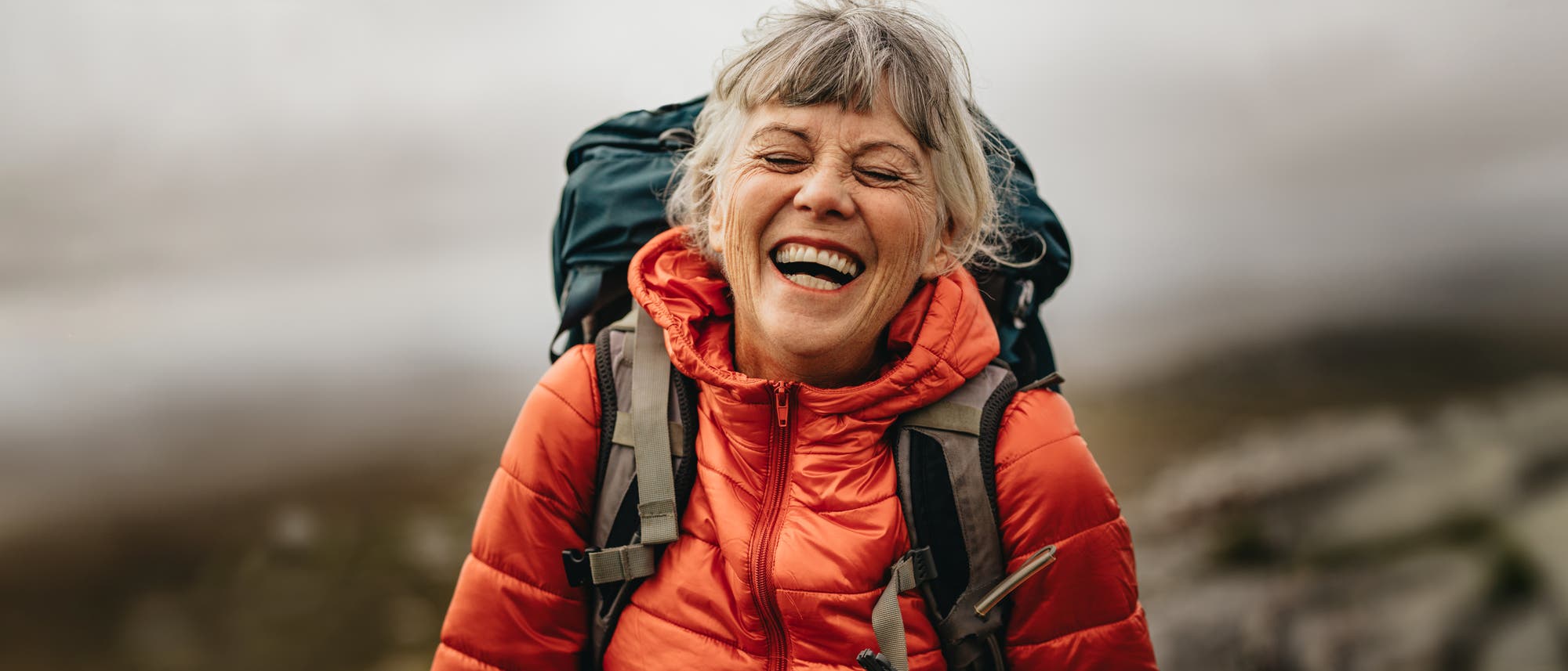 Eine ältere Frau mit grauem Haar lacht herzlich, während sie in einer roten Daunenjacke und mit einem großen Rucksack in einer nebligen, bergigen Landschaft wandert. Die Szene vermittelt Freude und Abenteuerlust. Eine ältere Frau mit grauem Haar lacht herzlich, während sie in einer roten Daunenjacke und mit einem großen Rucksack in einer nebligen, bergigen Landschaft wandert. Die Szene vermittelt Freude und Abenteuerlust.