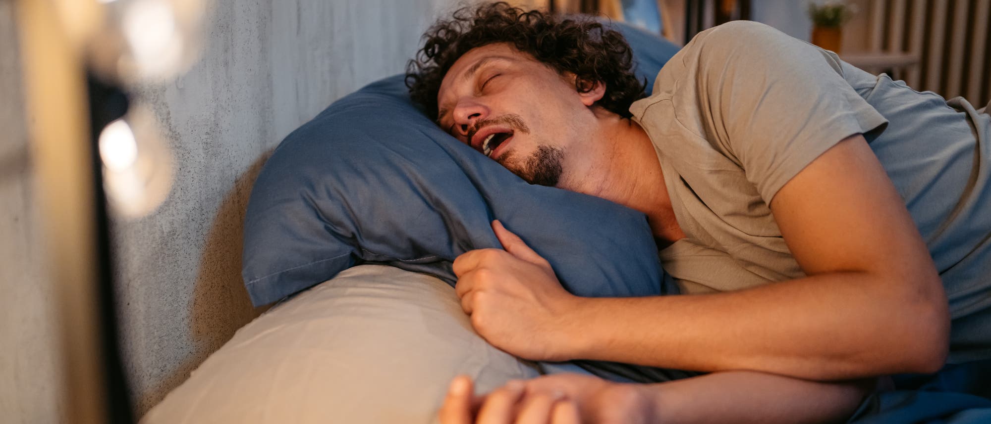 Eine Person liegt schlafend auf der Seite in einem Bett mit offenem Mund. Im Hintergrund ist eine sanfte Beleuchtung zu sehen.
