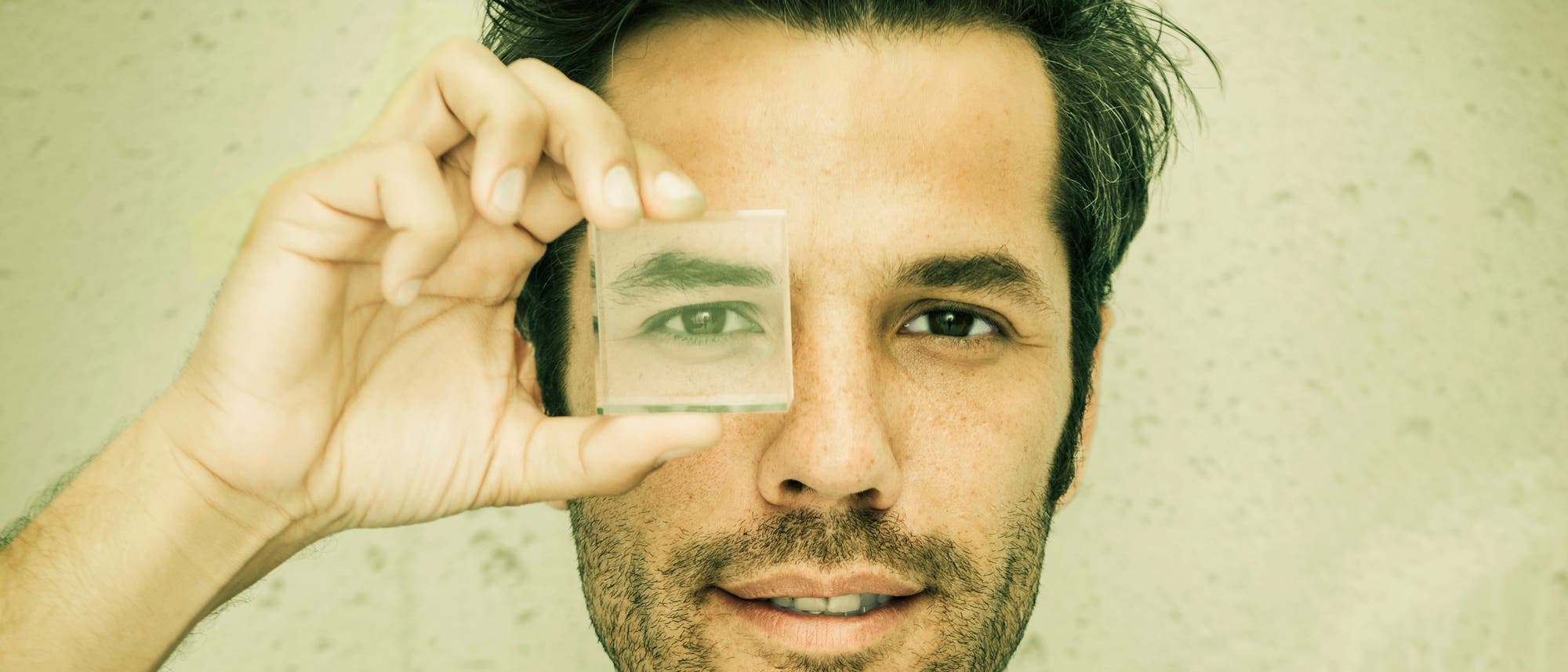 Eine Person h&auml;lt ein quadratisches Glasst&uuml;ck vor ihr rechtes Auge, wodurch das Auge vergr&ouml;&szlig;ert erscheint. Der Hintergrund ist eine einfarbige Wand. Die Person l&auml;chelt leicht und schaut direkt in die Kamera.